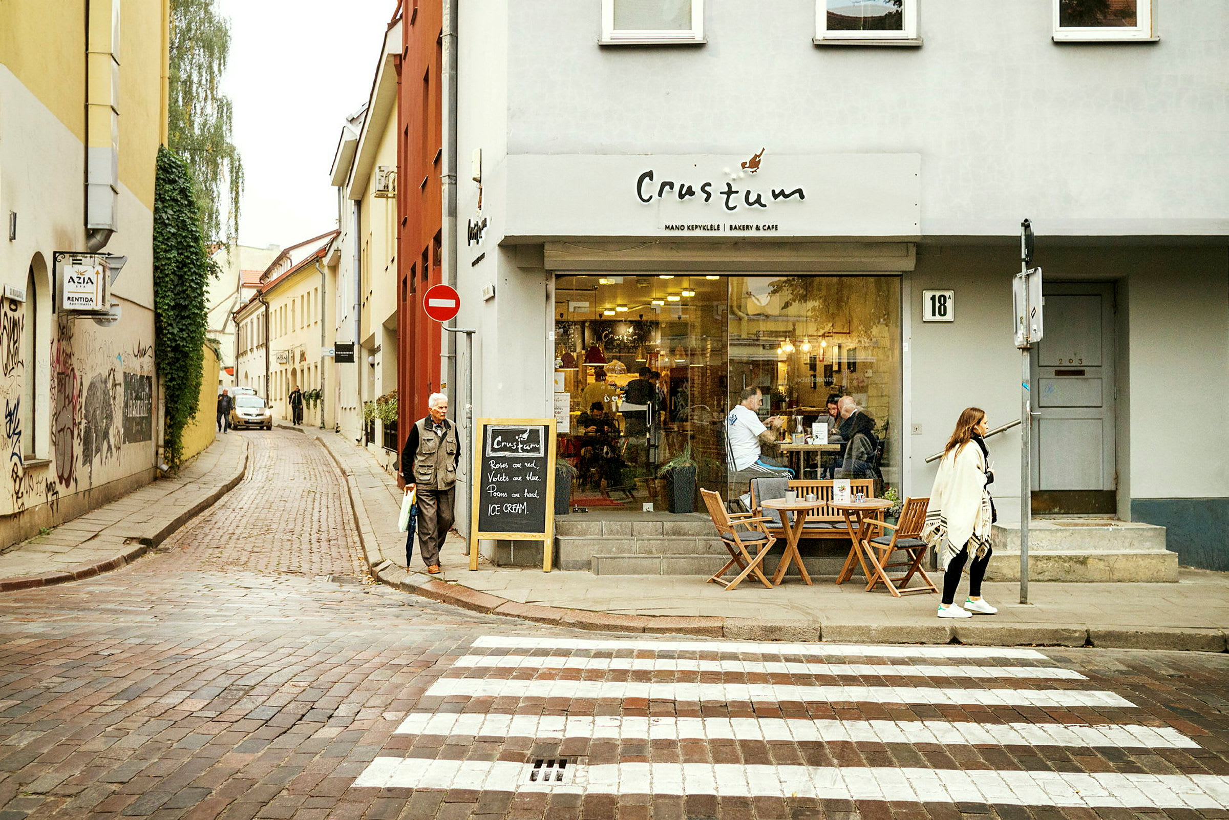 En gade med en café på højre side. Folk sidder udendørs på caféens udendørsservering, og en person krydser et fodgængerfelt. (AI-genereret tekst)