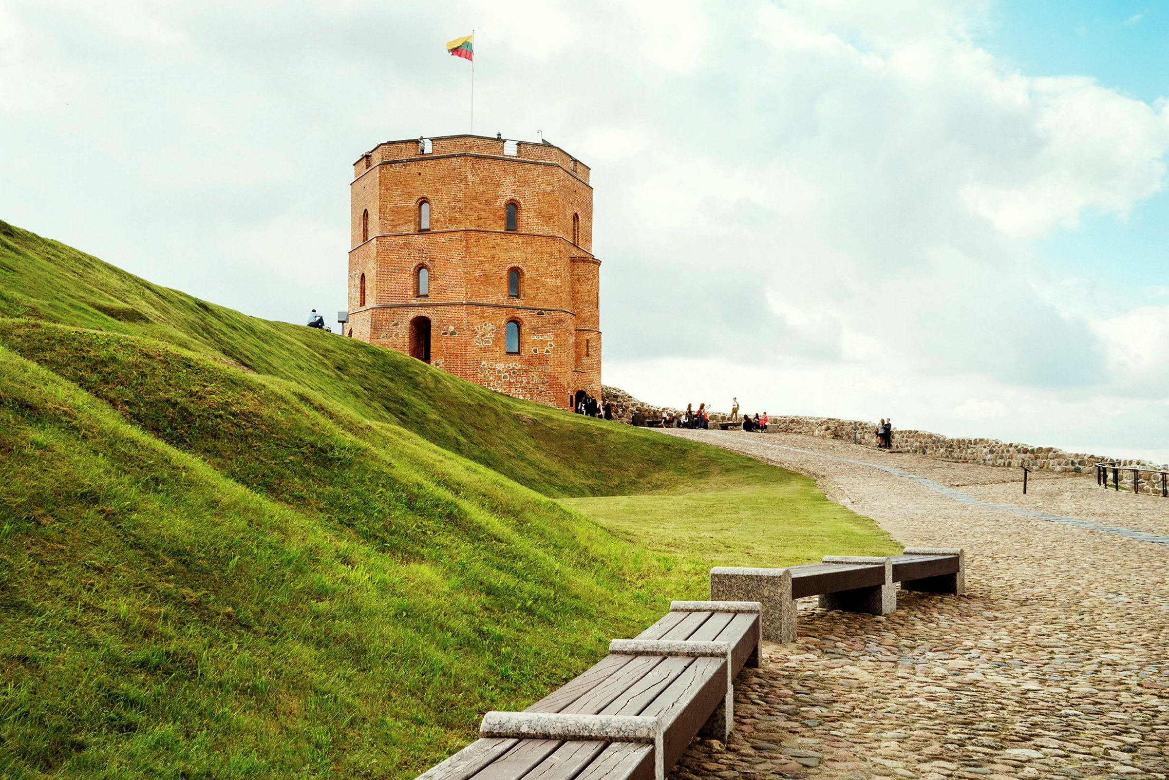 En gammel teglstensbygning med et rundt tårn står på en bakke med grønt græs. På tårnet vajer et litauisk flag. (AI-genereret tekst)