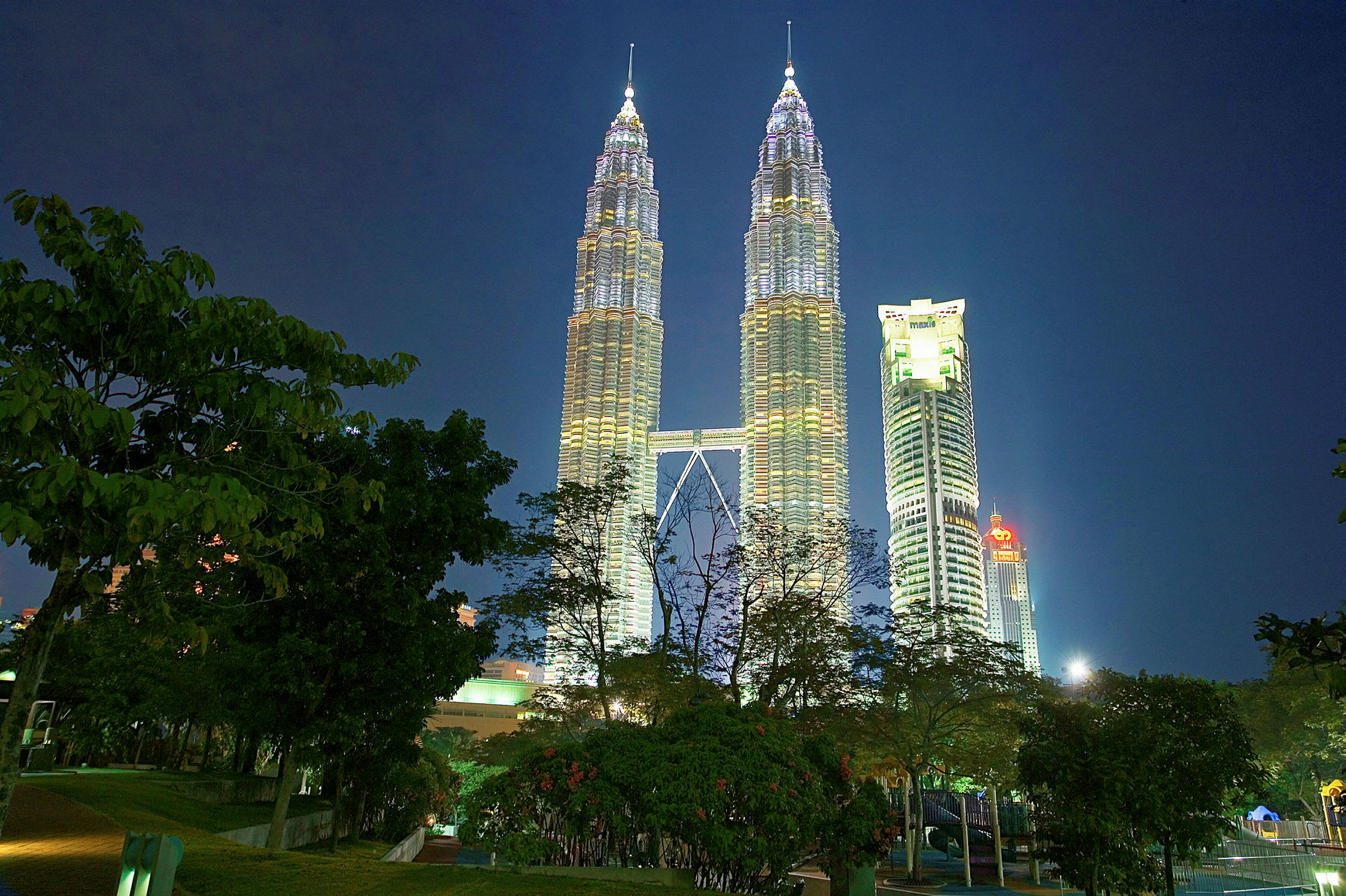 Petronas Twin Towers oplyst om natten med træer i forgrunden. (AI-genereret tekst)