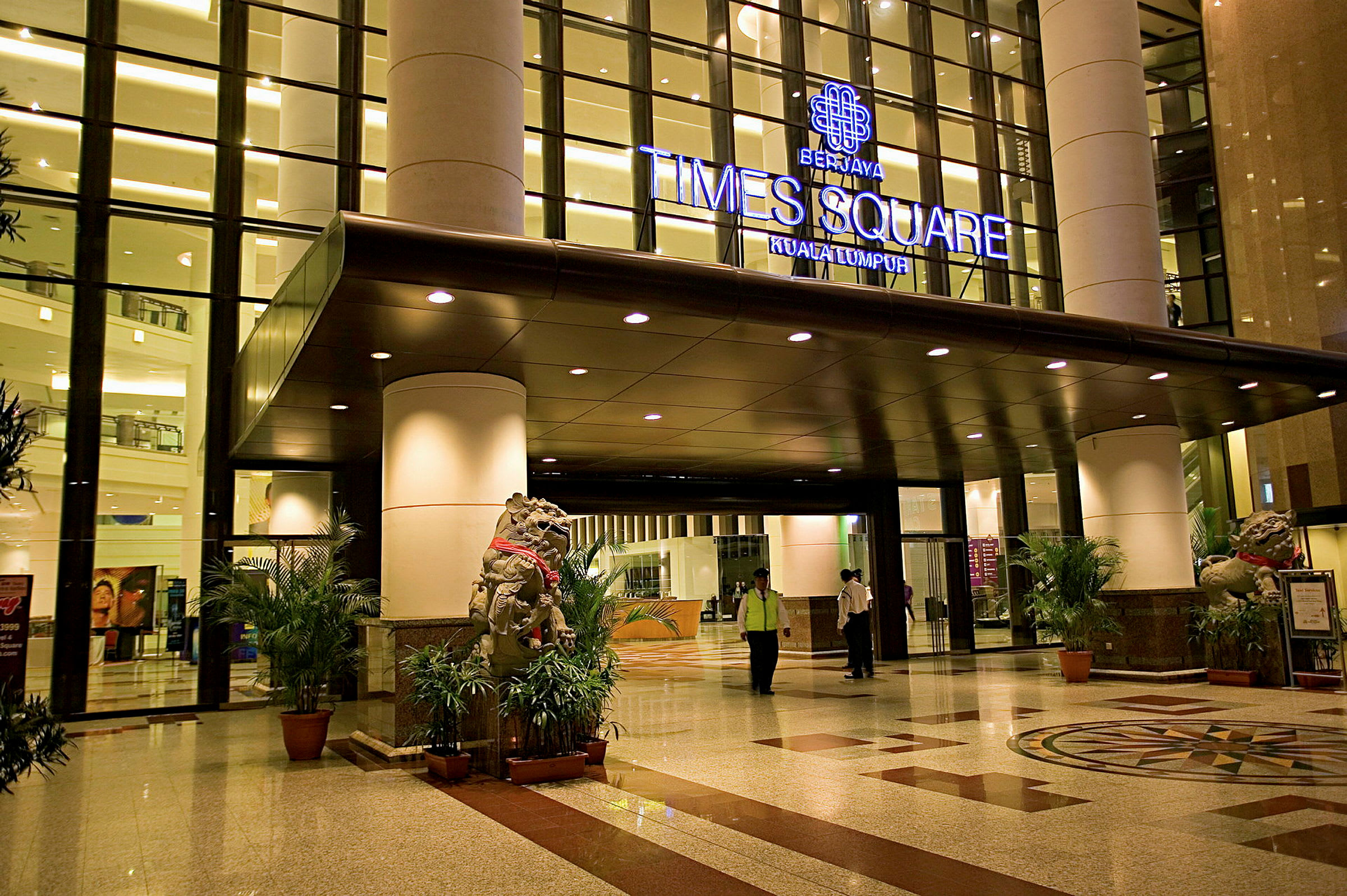 Indgangsområde til en bygning med glasfacade og lys belysning. Et skilt over indgangen angiver 'Berjaya Times Square Kuala Lumpur'. Trægulv og dekorative planter er også synlige. (AI-genereret tekst)