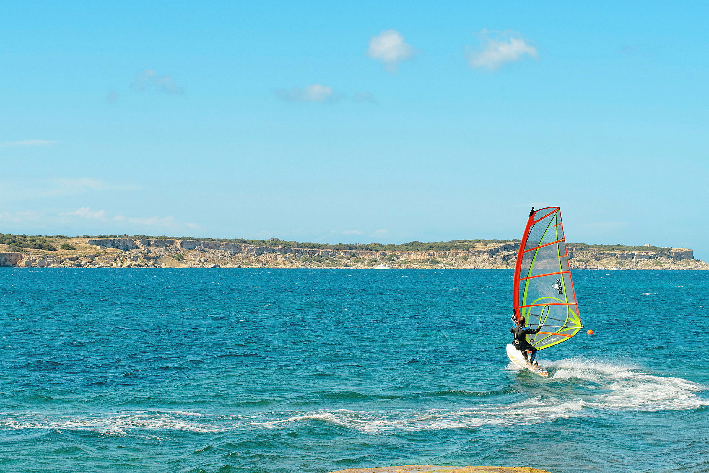 Person, der windsurfer på en blå havudsigt med klipper i horisonten. (AI-genereret tekst)
