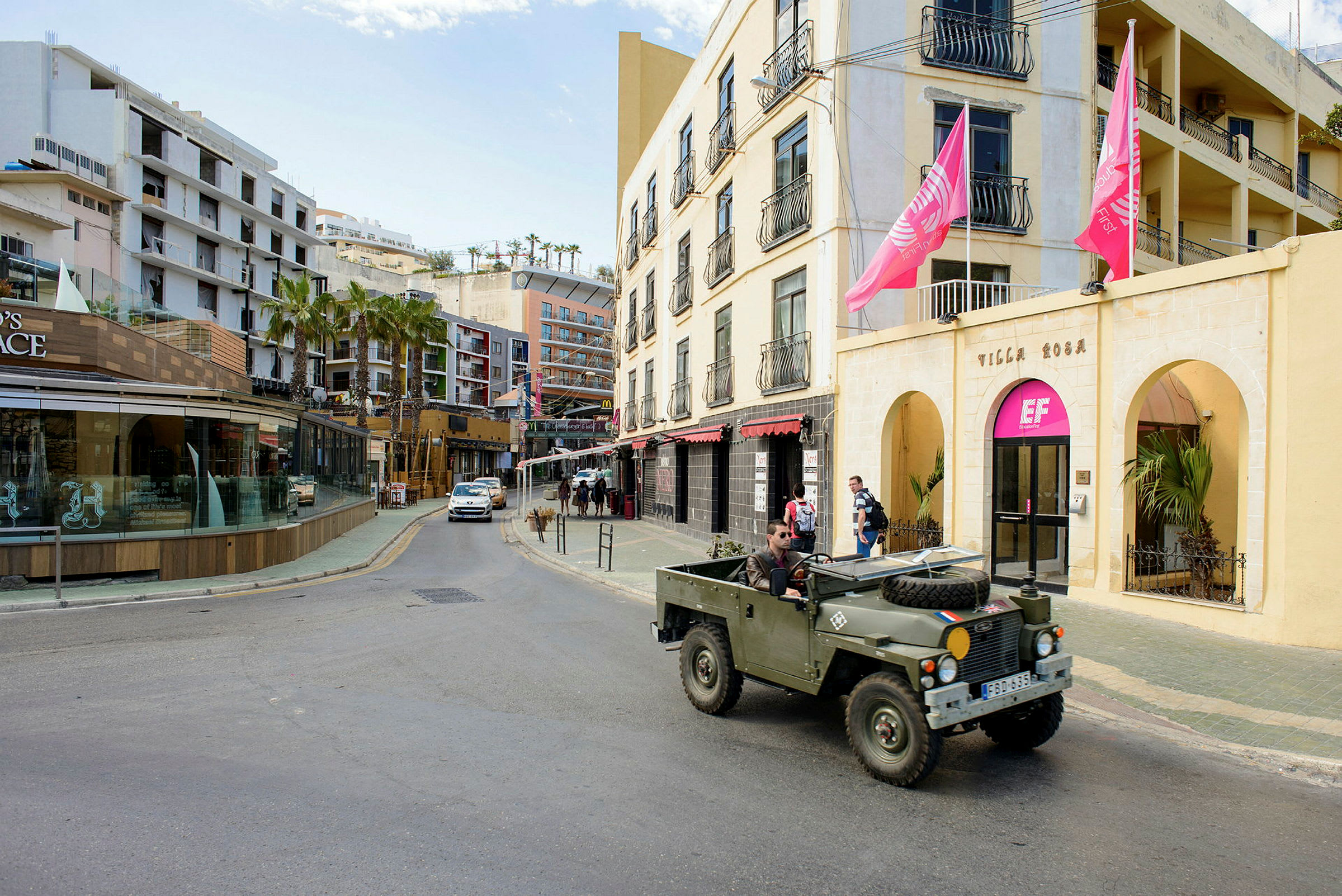 En gade med bygninger, herunder en gul facade med lyserøde flag og en butik med glasvinduer. En militærgrøn jeep er parkeret ved vejkanten. (AI-genereret tekst)