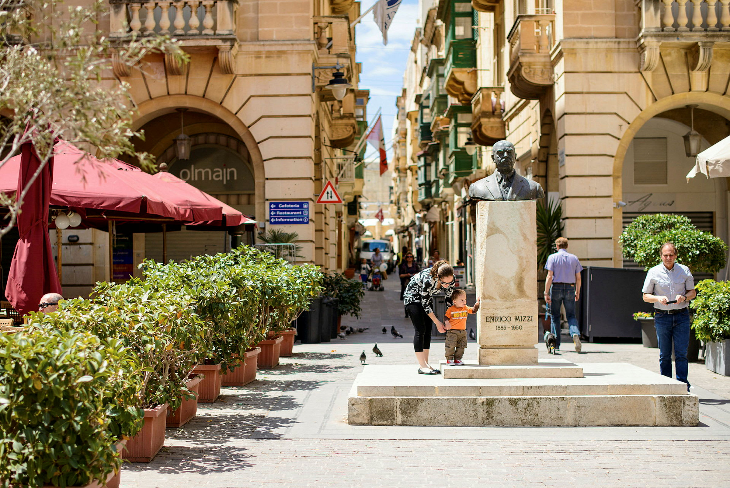 En gade med statuen af Enrico Mizzi, omgivet af bygninger og beplantning. (AI-genereret tekst)