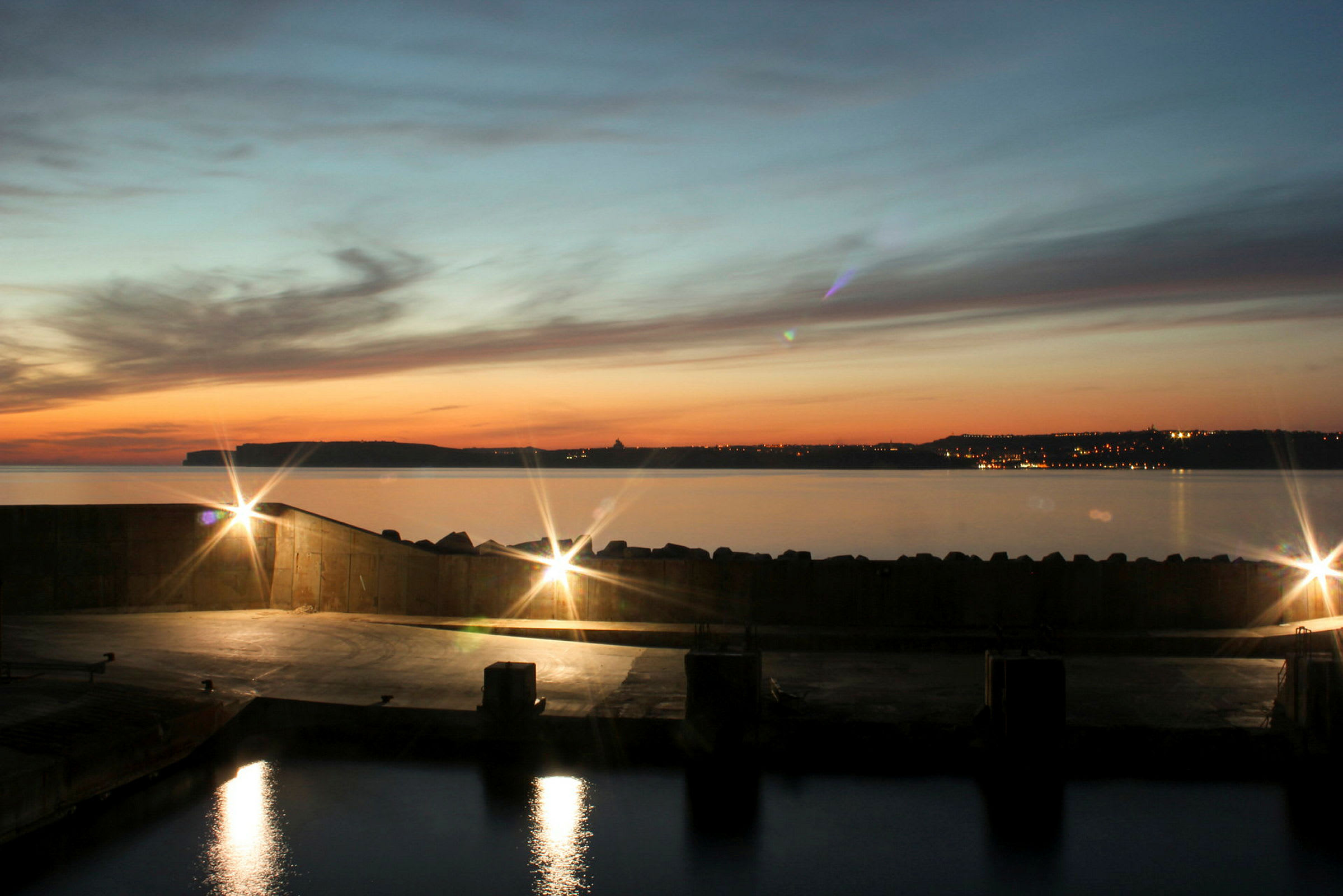 Havnebassin med lysreflekser på vandet, kysten og byens lys er synlige ved solnedgang. (AI-genereret tekst)