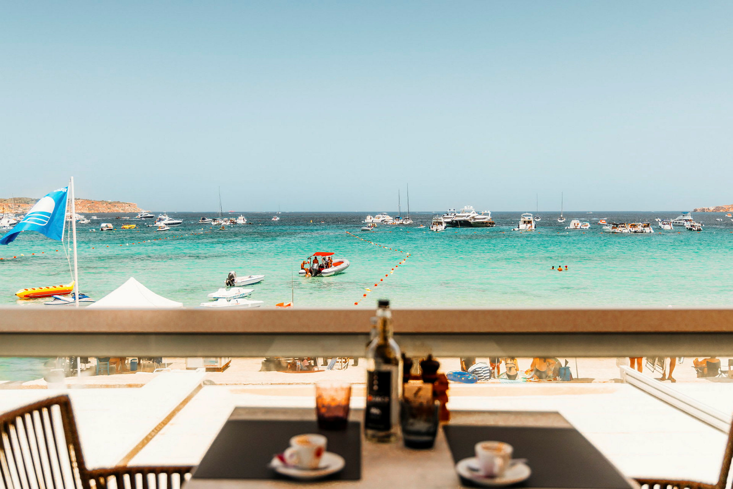 Udsigt fra en udendørs restaurant mod en strand med mennesker, havet og små både. (AI-genereret tekst)