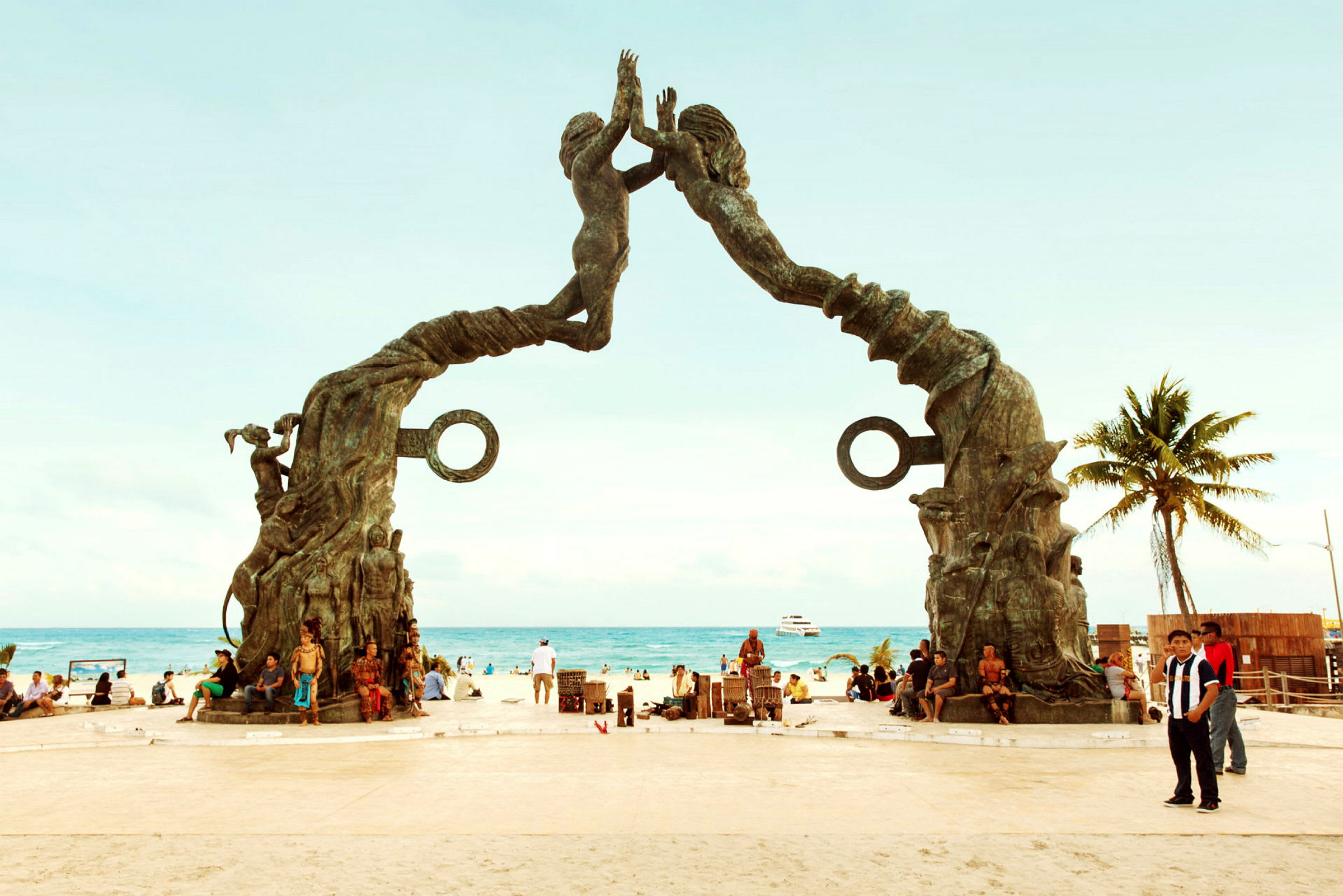 Et monument på stranden med to stenstatuer, der danner en bue. Bag monumentet kan man se havet og mennesker på stranden. (AI-genereret tekst)