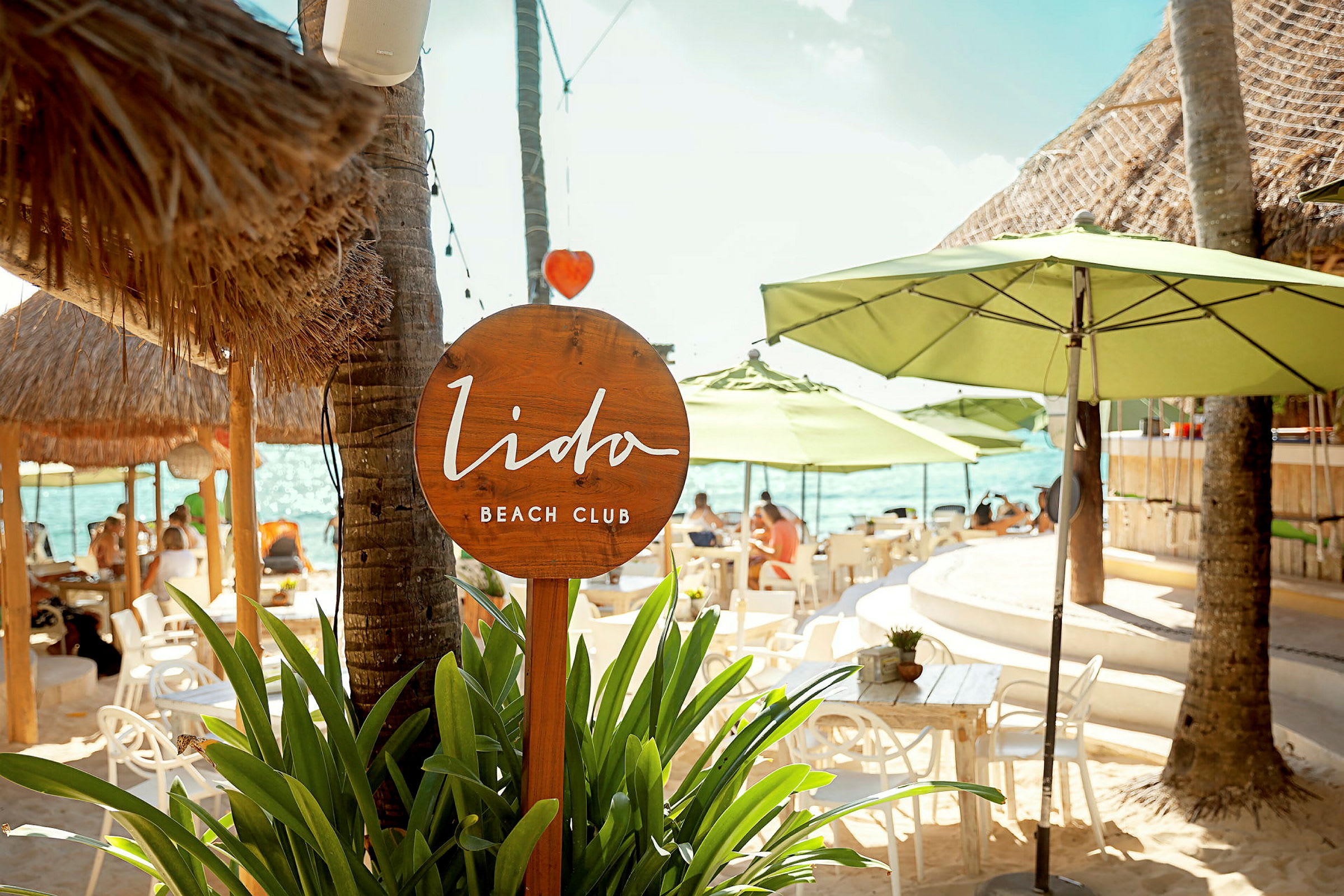Et skilt for Lido Beach Club omgivet af grønne planter og parasoller på en strand. (AI-genereret tekst)