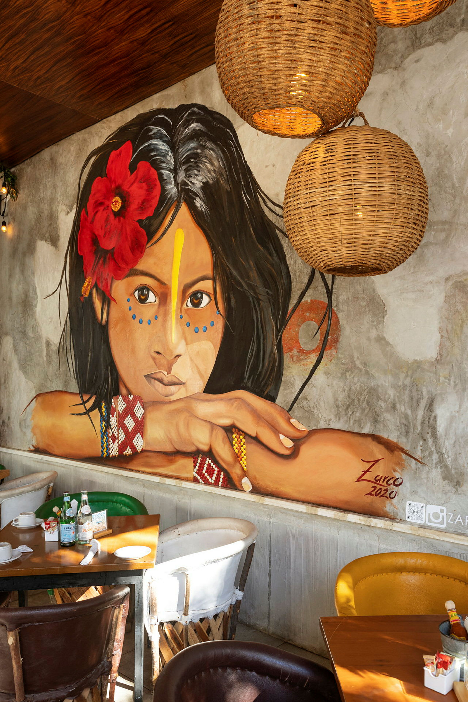 Et vægmaleri af en ung kvinde med en rød blomst i håret og farverige mønstre, omgivet af interiør i en restaurant. (AI-genereret tekst)