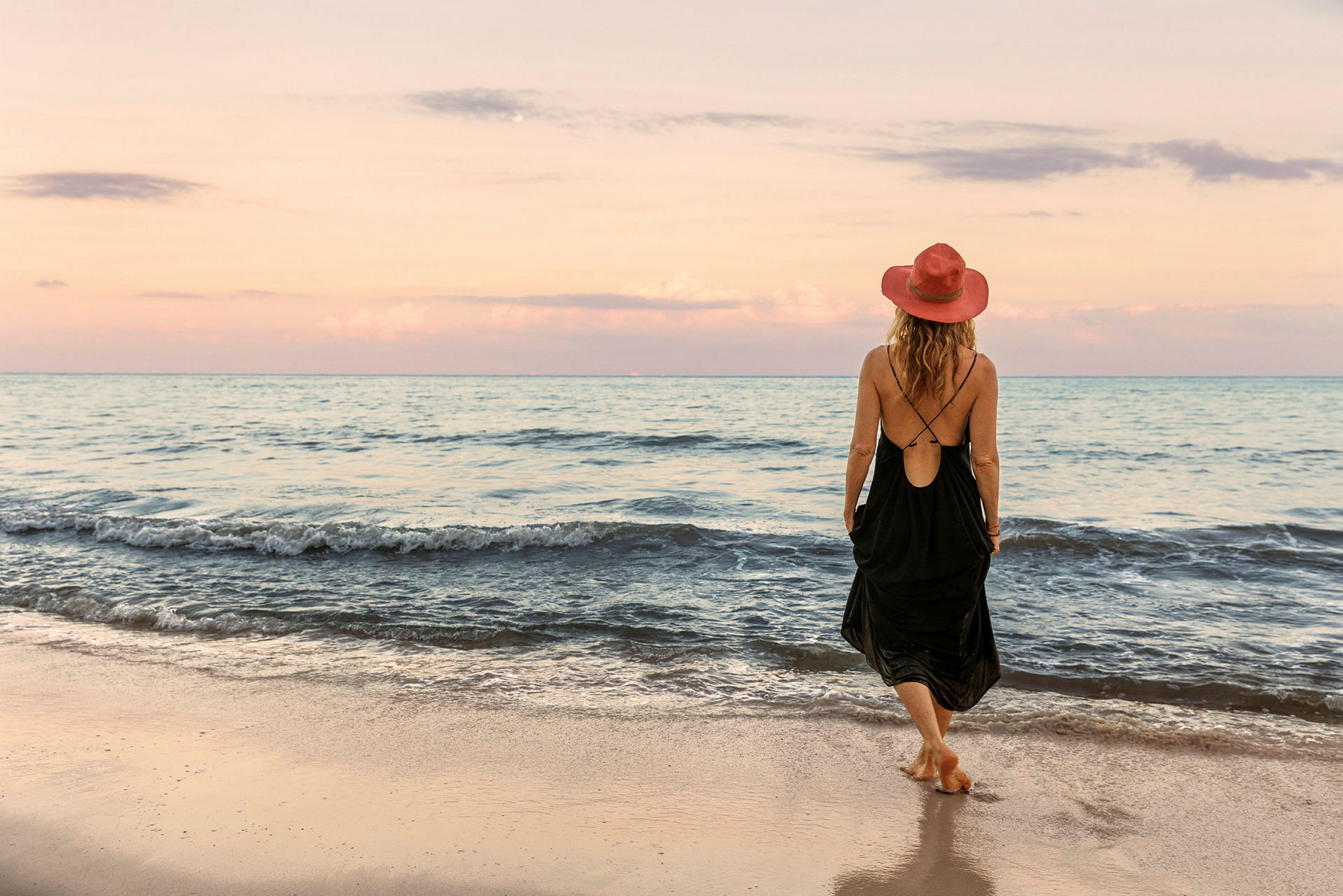 En person med en rød hat og sort kjole går på en sandstrand ved havet under en pastelfarvet himmel. (AI-genereret tekst)