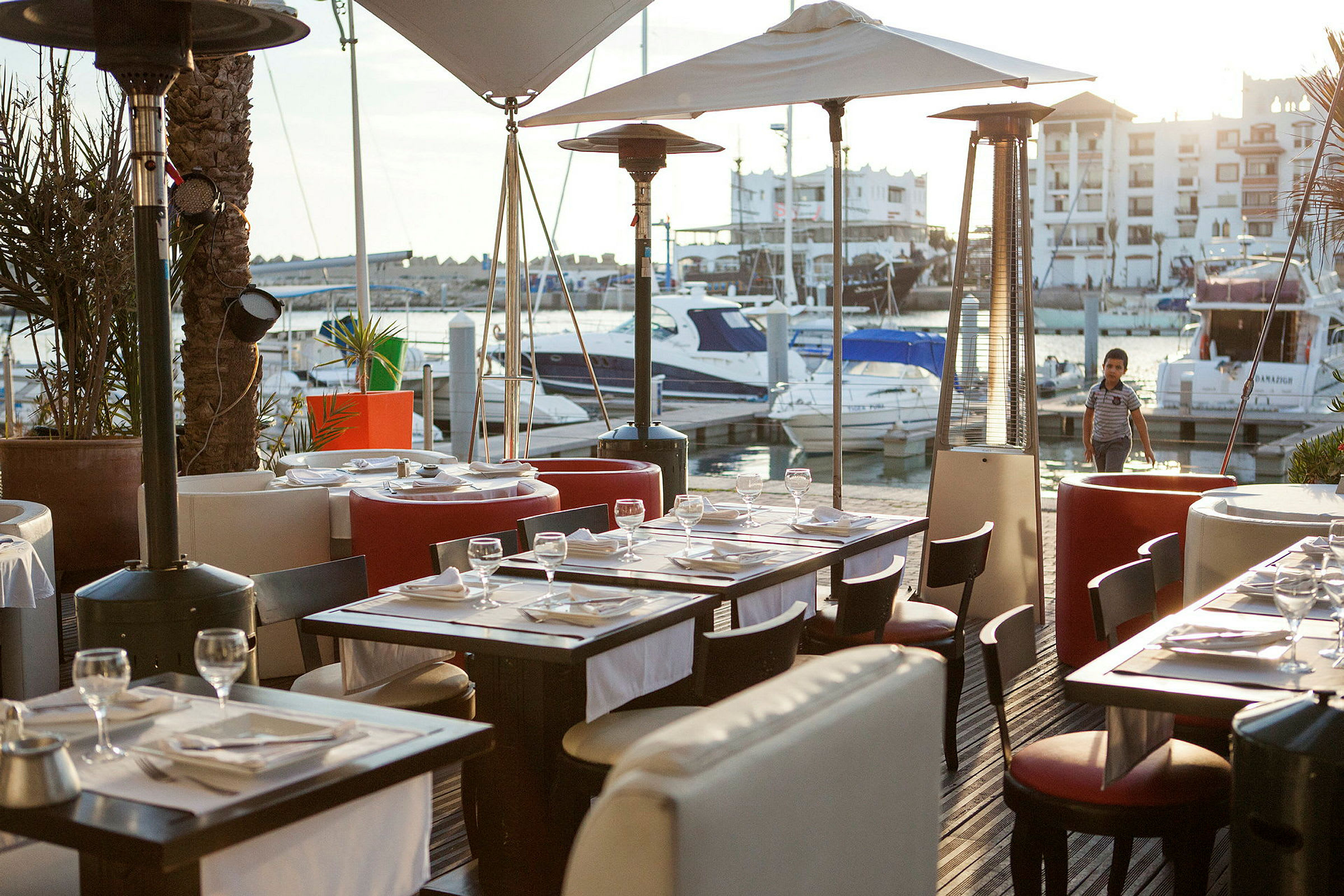 Udendørs restaurant ved en marina med både og bygninger i baggrunden. (AI-genereret tekst)
