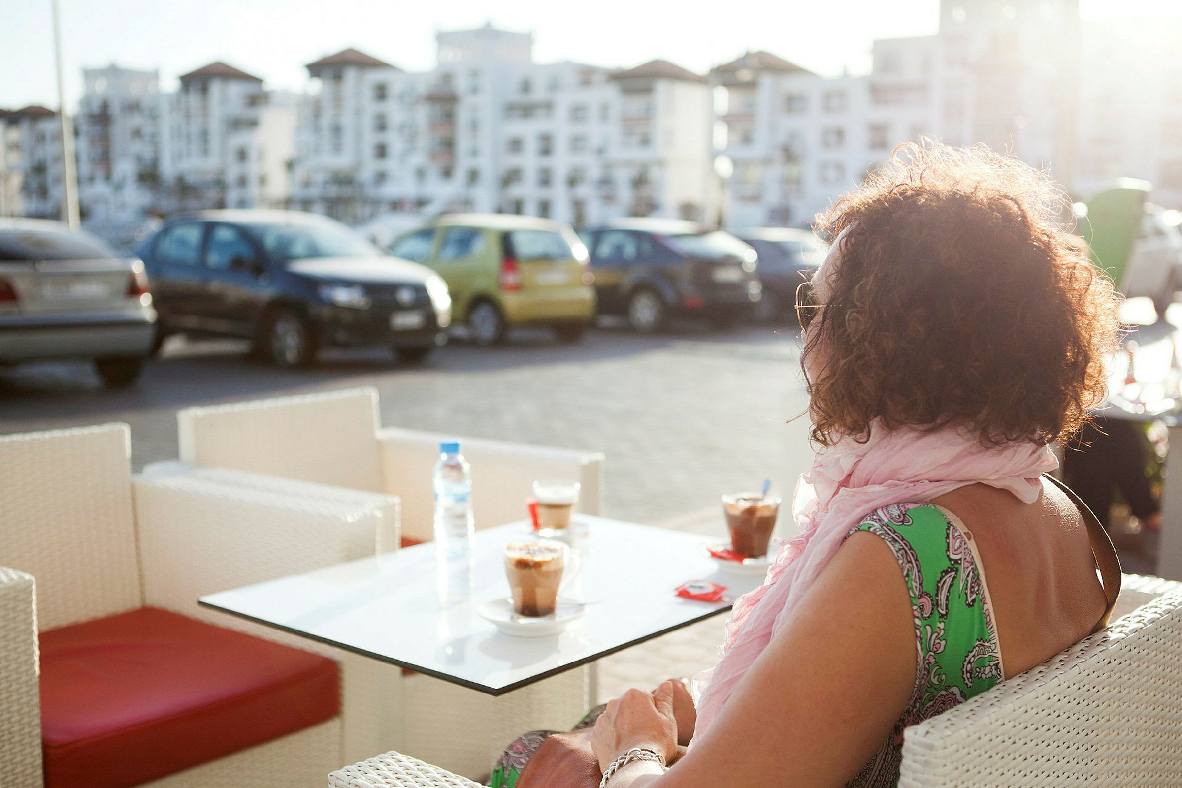 En person sidder ved et bord udendørs med to kaffedrikke, i baggrunden ses parkerede biler og bygninger. (AI-genereret tekst)