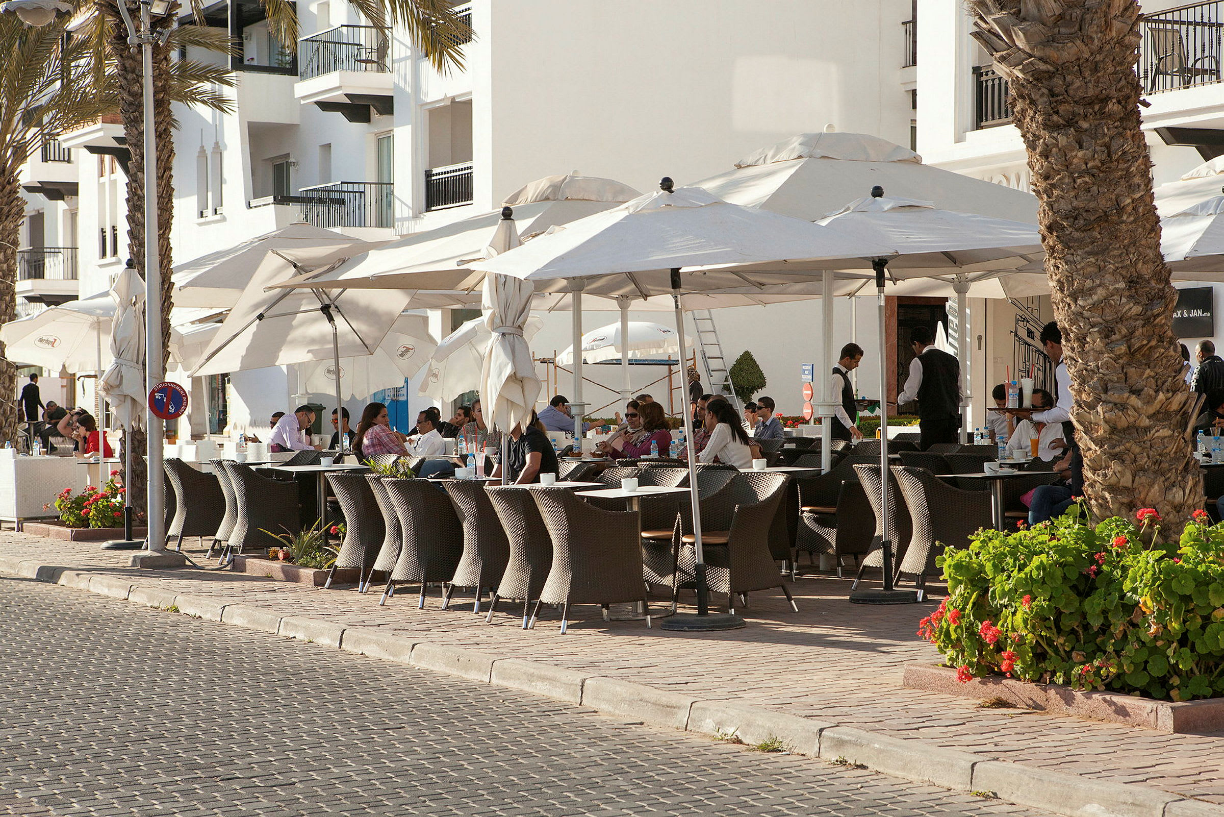 Udendørs restaurantområde med borde, stole og parasoller ved en brostensbelagt gangsti. (AI-genereret tekst)