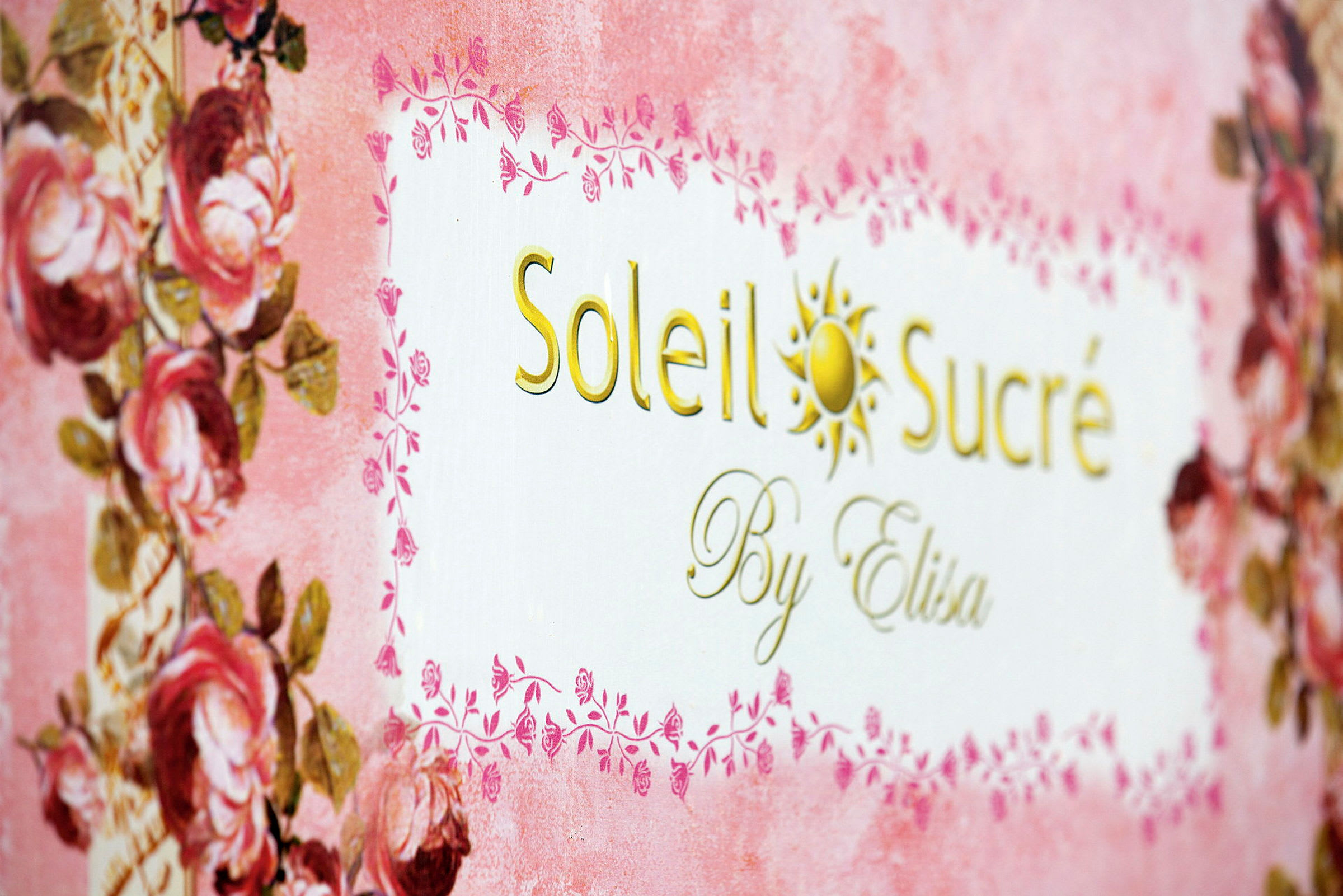 Teksten 'Soleil Sucré By Elisa' på en dekoreret baggrund med lyserøde og blomsterdetaljer. (AI-genereret tekst)