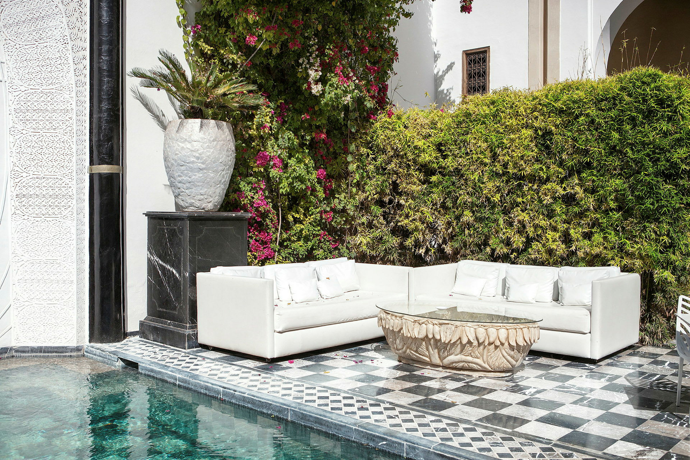 En terrasse med en hvid sofa, et udsmykket bord og en pool ved siden af en væg med klatreplanter og blomster. (AI-genereret tekst)