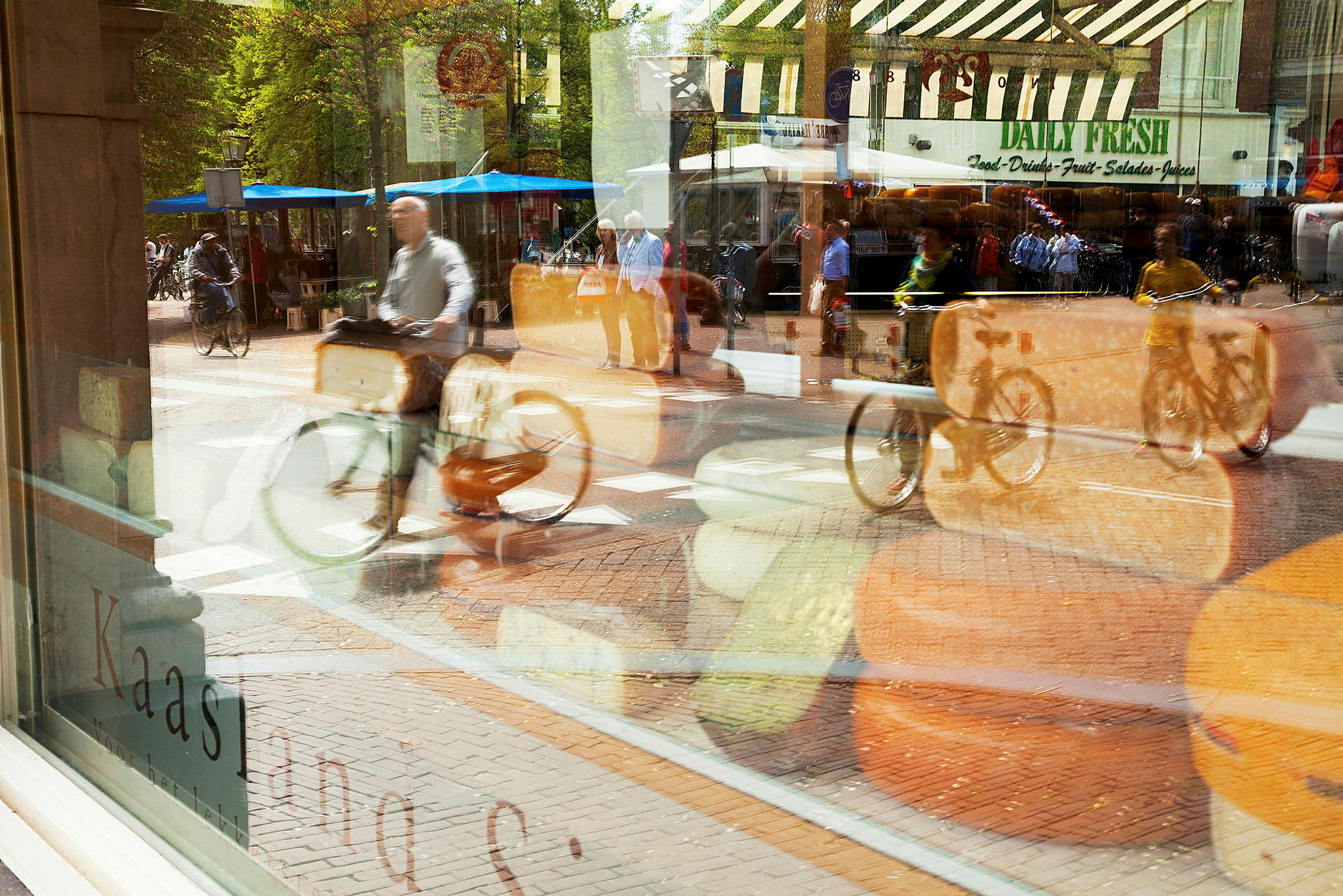 Et butiksvindue med ost og en refleksion af en gade med cyklister og markedsboder ses. (AI-genereret tekst)