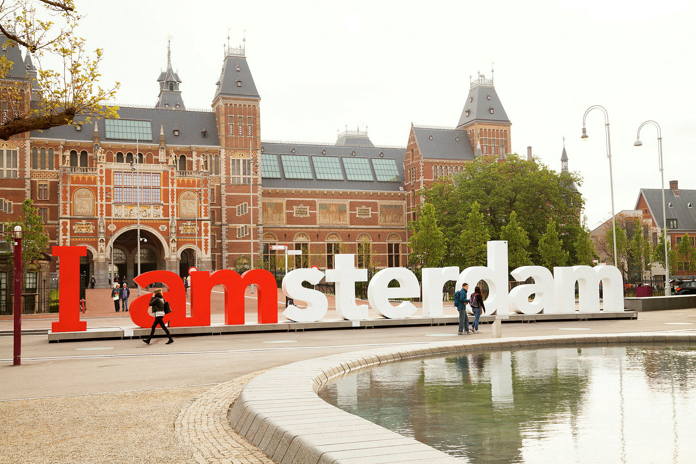 Stor bygning i mursten med flere tårne bag et I amsterdam-skilt ved vandet. (AI-genereret tekst)