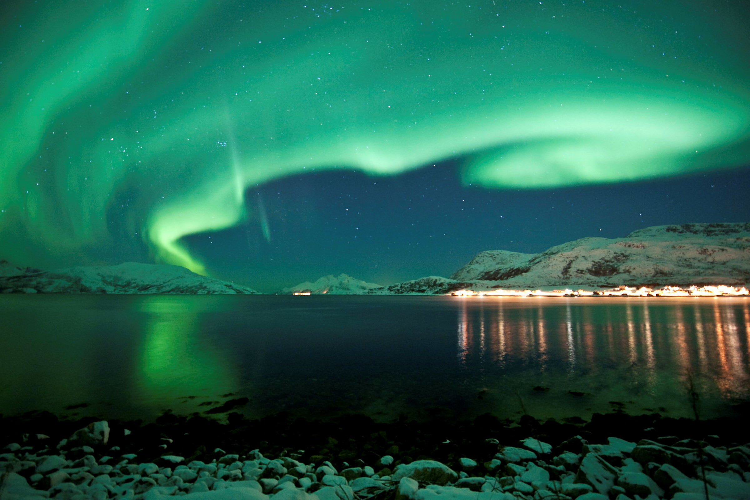 Billede af et naturfænomen, der viser nordlys over et landskab med sneklædte bjerge, vand og en kystlinje med lys fra bygninger. (AI-genereret tekst)