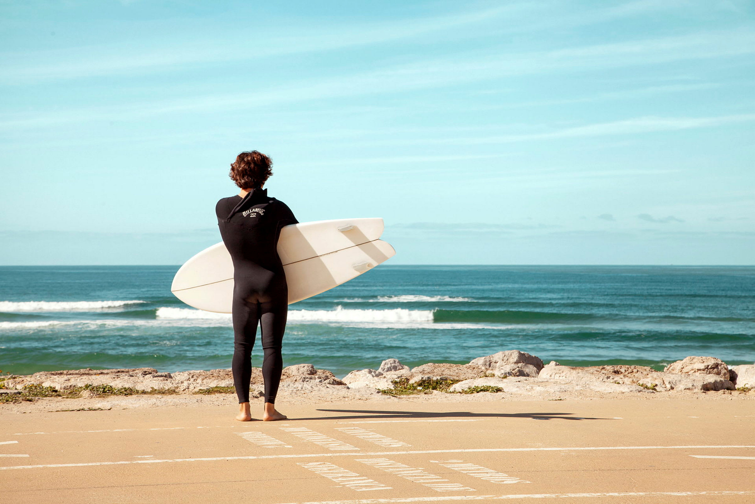 En person i våddragt holder et surfbræt og står vendt mod havet. (AI-genereret tekst)