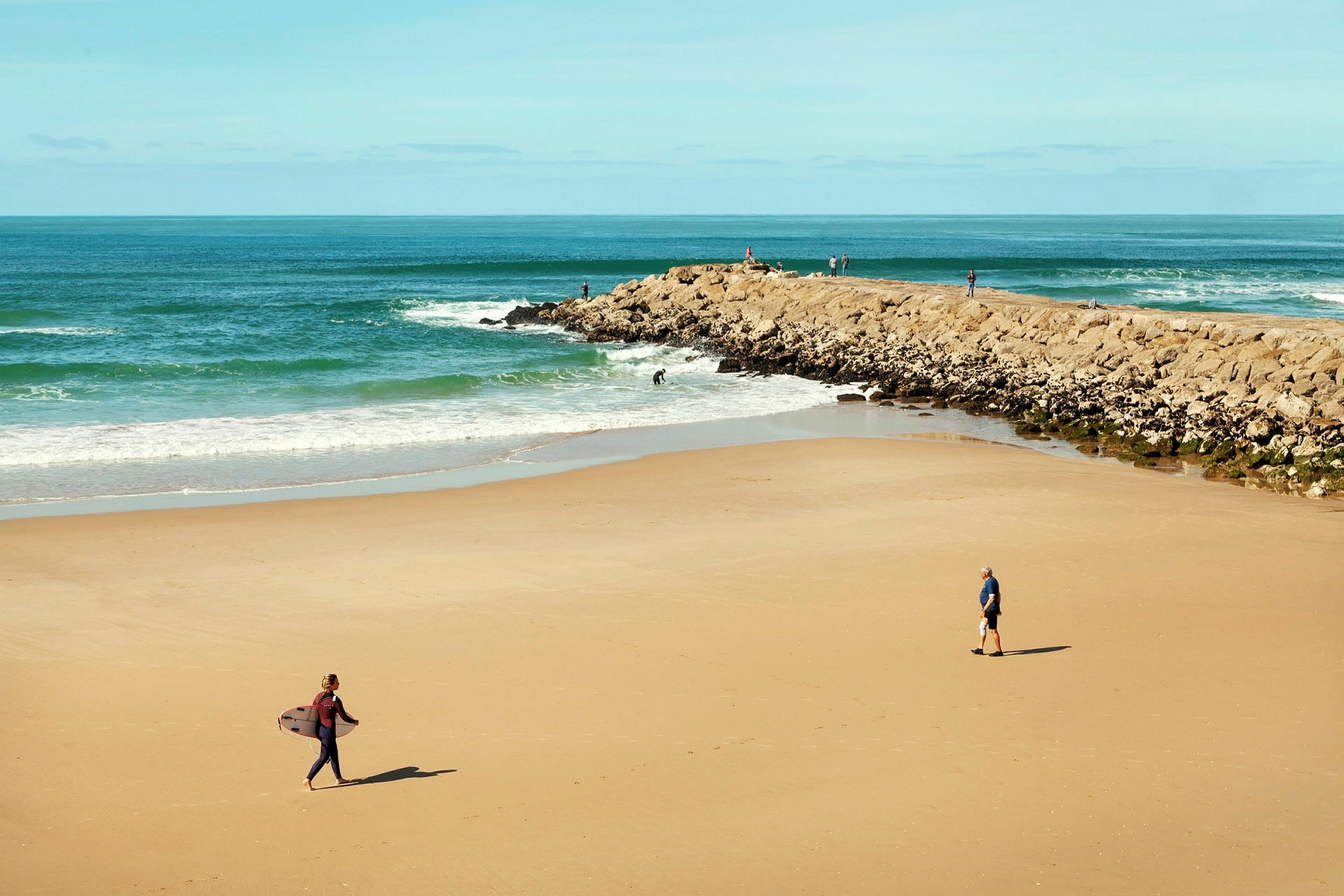 En strand med nogle personer, hvoraf en af dem bærer et surfbræt. (AI-genereret tekst)