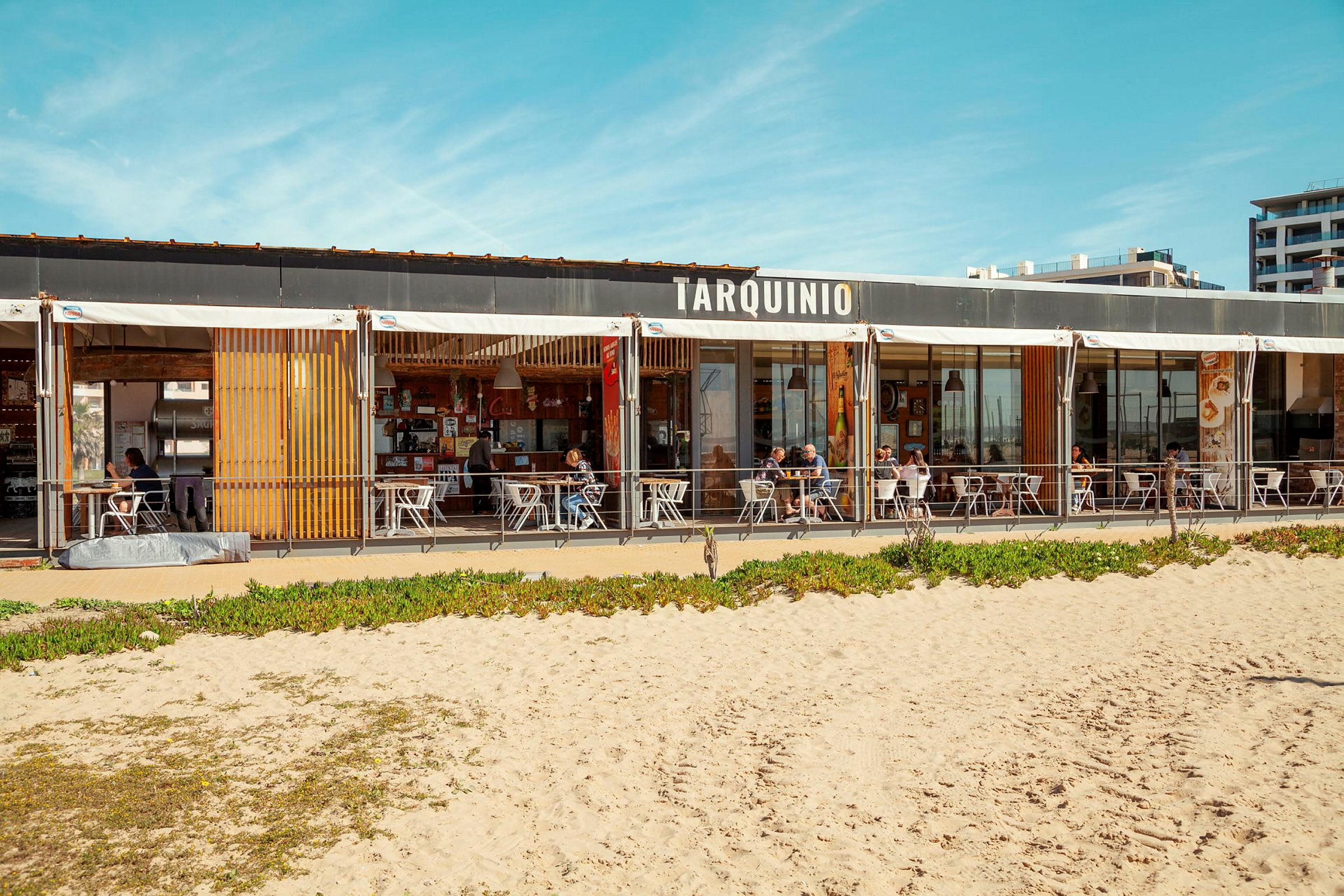 En strandrestaurant med skilt, der siger 'Tarquinio', siddepladser udendørs og sandstrand i forgrunden. (AI-genereret tekst)