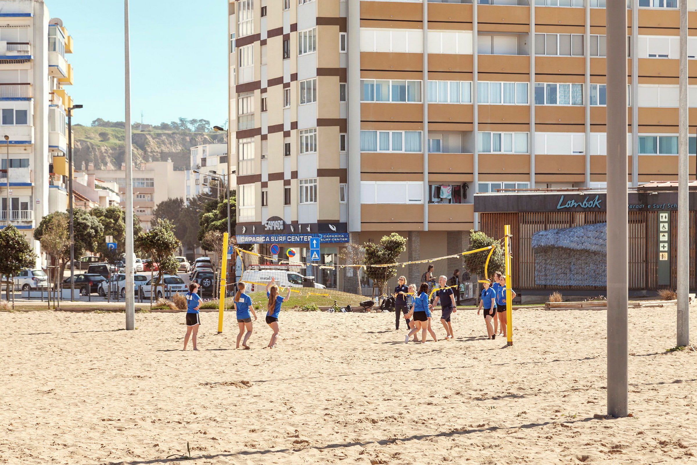 En gruppe mennesker spiller volleyball på en sandbane med bygninger i baggrunden. (AI-genereret tekst)