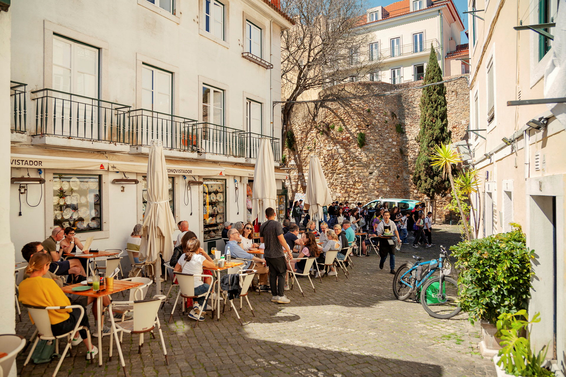 En udendørsterrasse ved en restaurant med flere mennesker, der sidder ved borde og nyder deres måltid. I baggrunden ses en stenmur og en træbevokset bygning. (AI-genereret tekst)