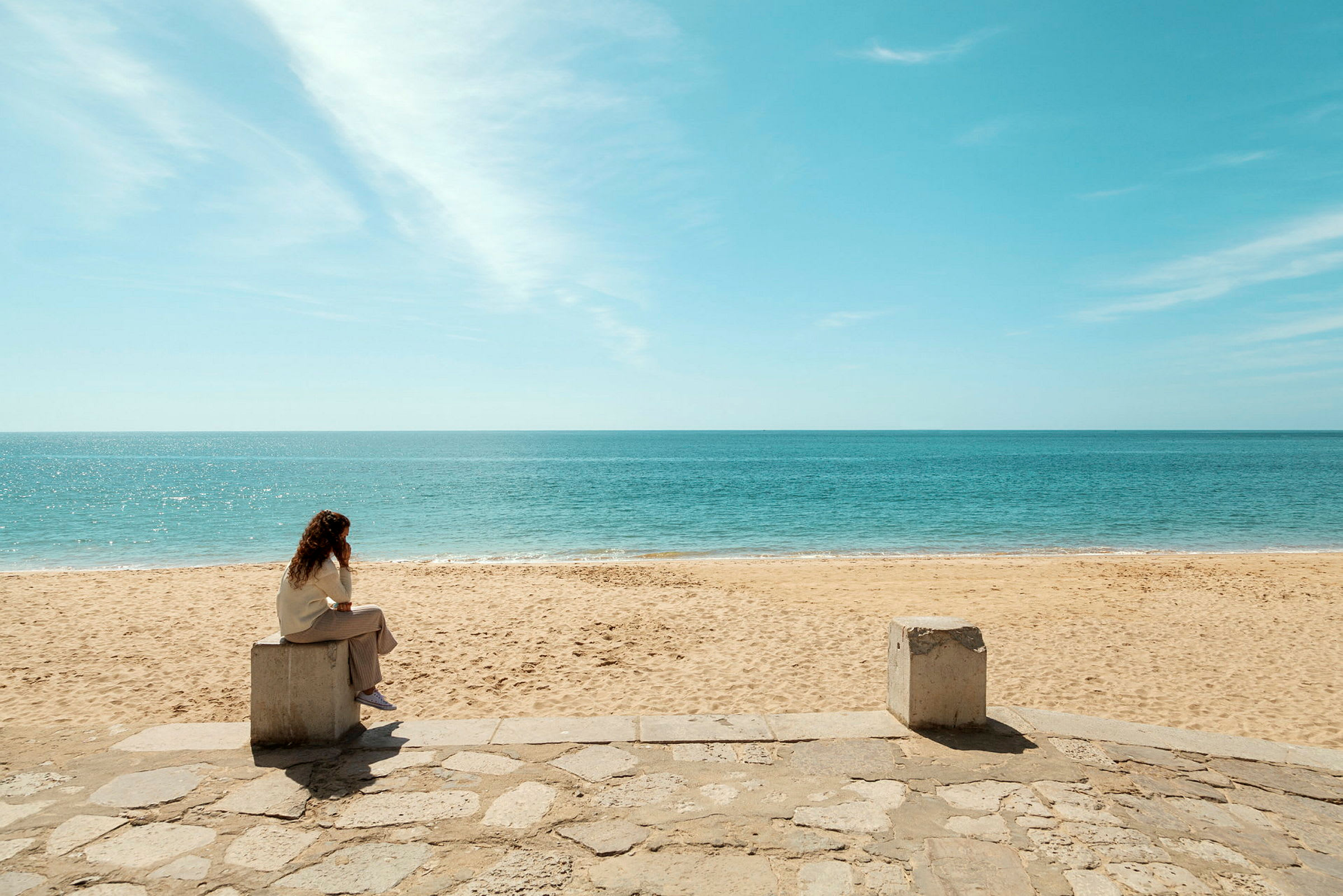 En person sidder på en betonblok ved en sandstrand med udsigt over havet. (AI-genereret tekst)