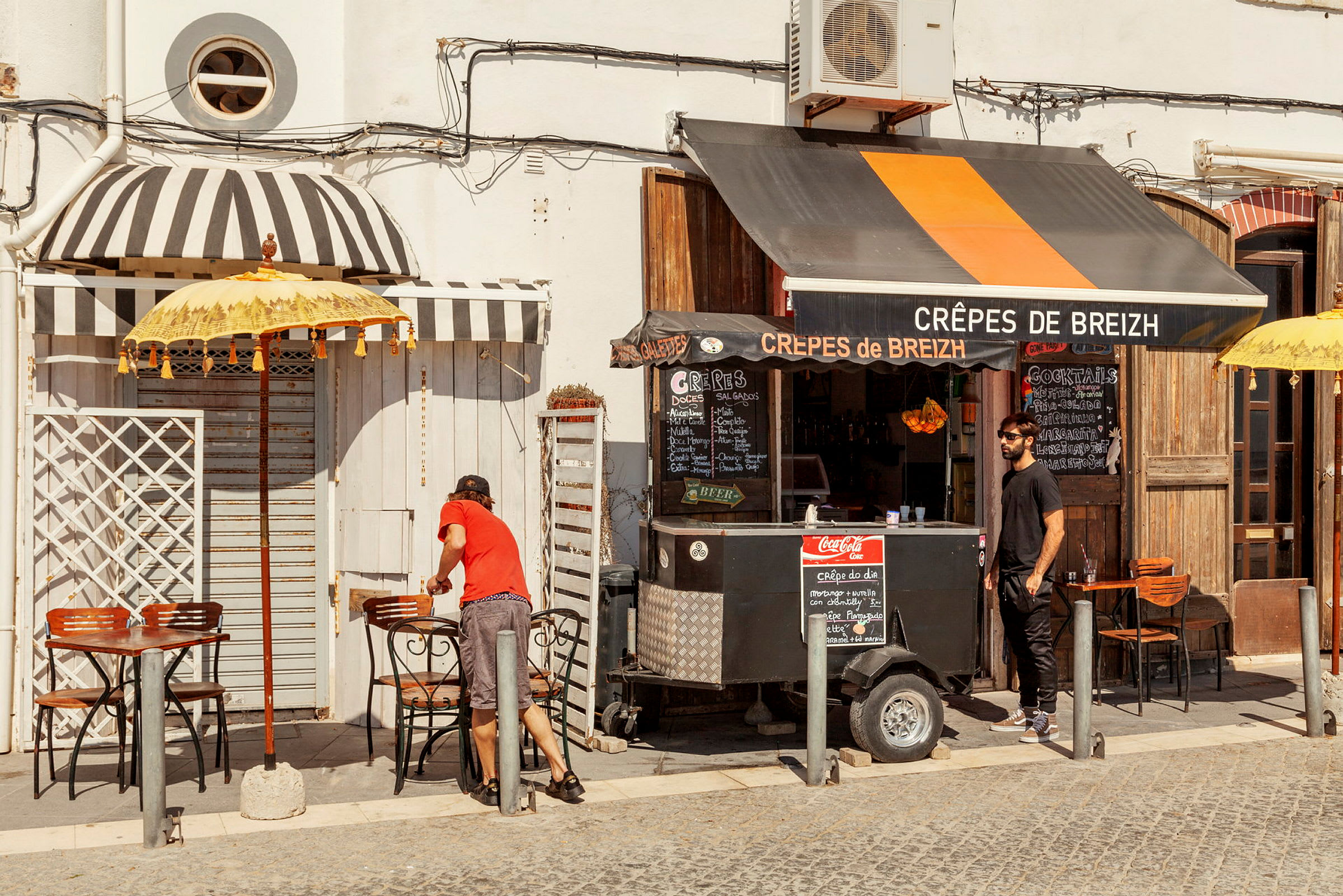 En lille madvogn, der sælger crêpes, med borde og siddepladser udendørs på en solrig dag. (AI-genereret tekst)