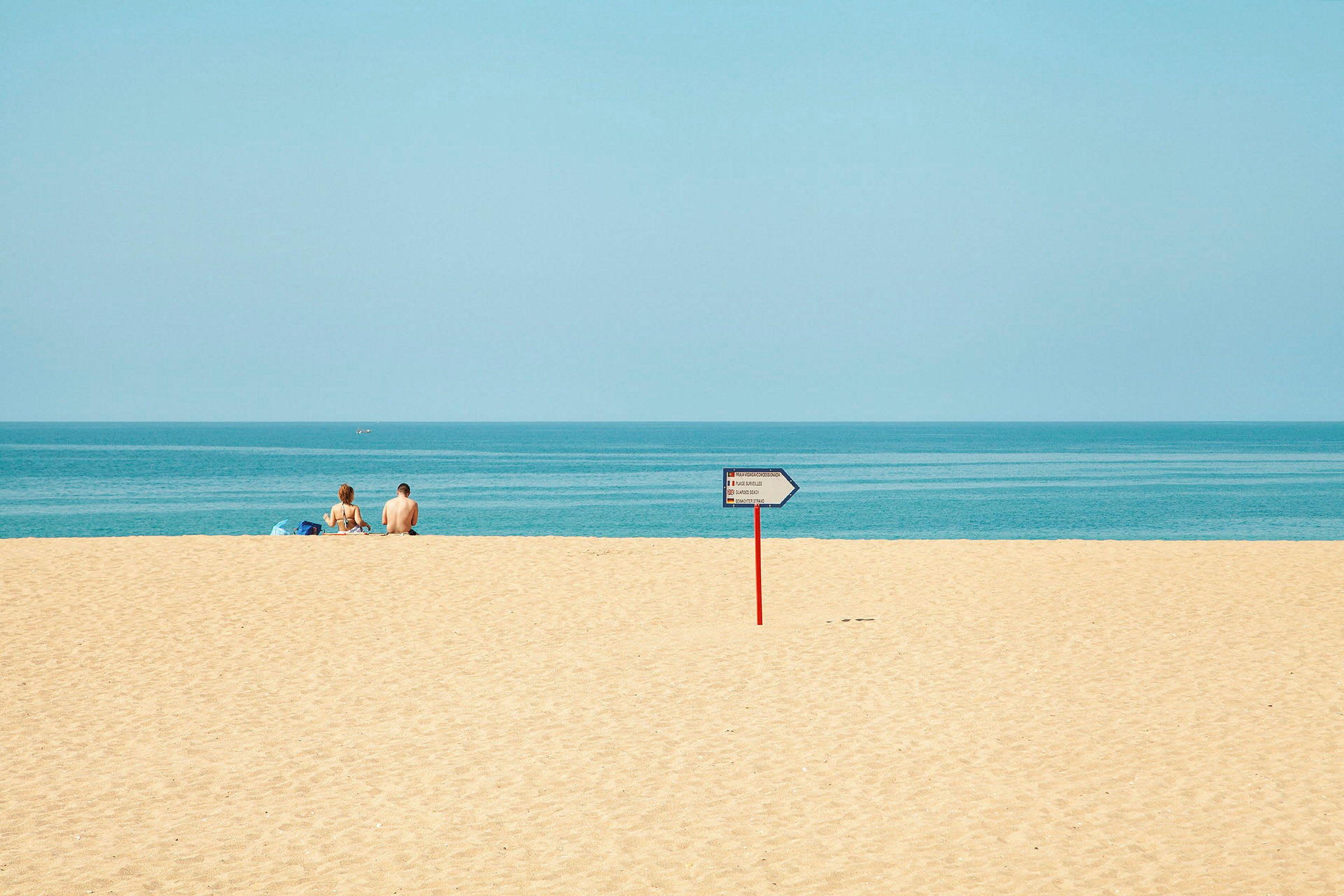 To personer sidder på en sandstrand med havet i baggrunden, og et vejskilt er synligt på stranden. (AI-genereret tekst)