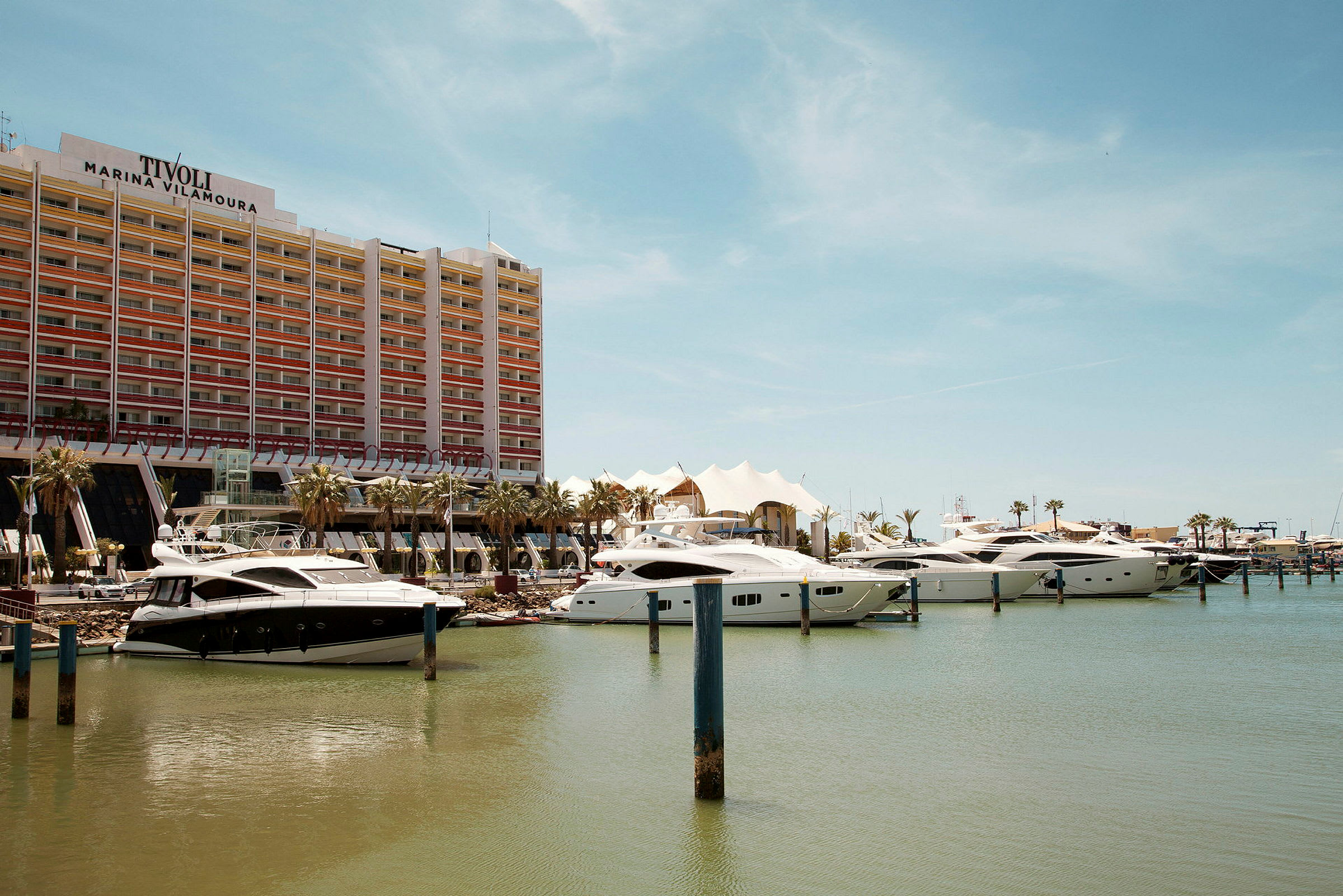 En moderne bygning med Tivoli Marina Vilamoura-logoen bag tilkoblede lystbåde ved en havn. (AI-genereret tekst)