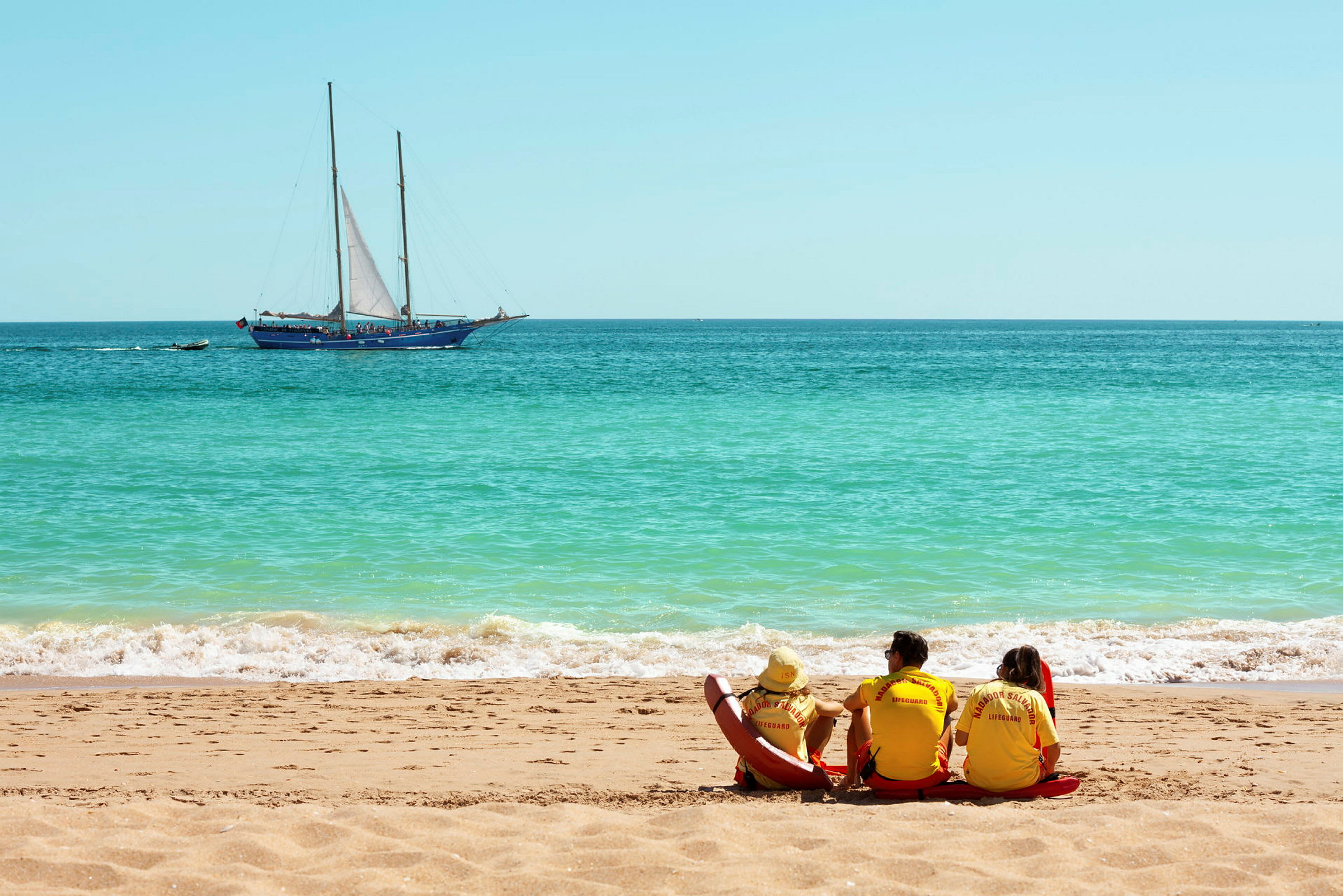 Livreddere sidder på en strand med en sejlbåd på havet i baggrunden. (AI-genereret tekst)