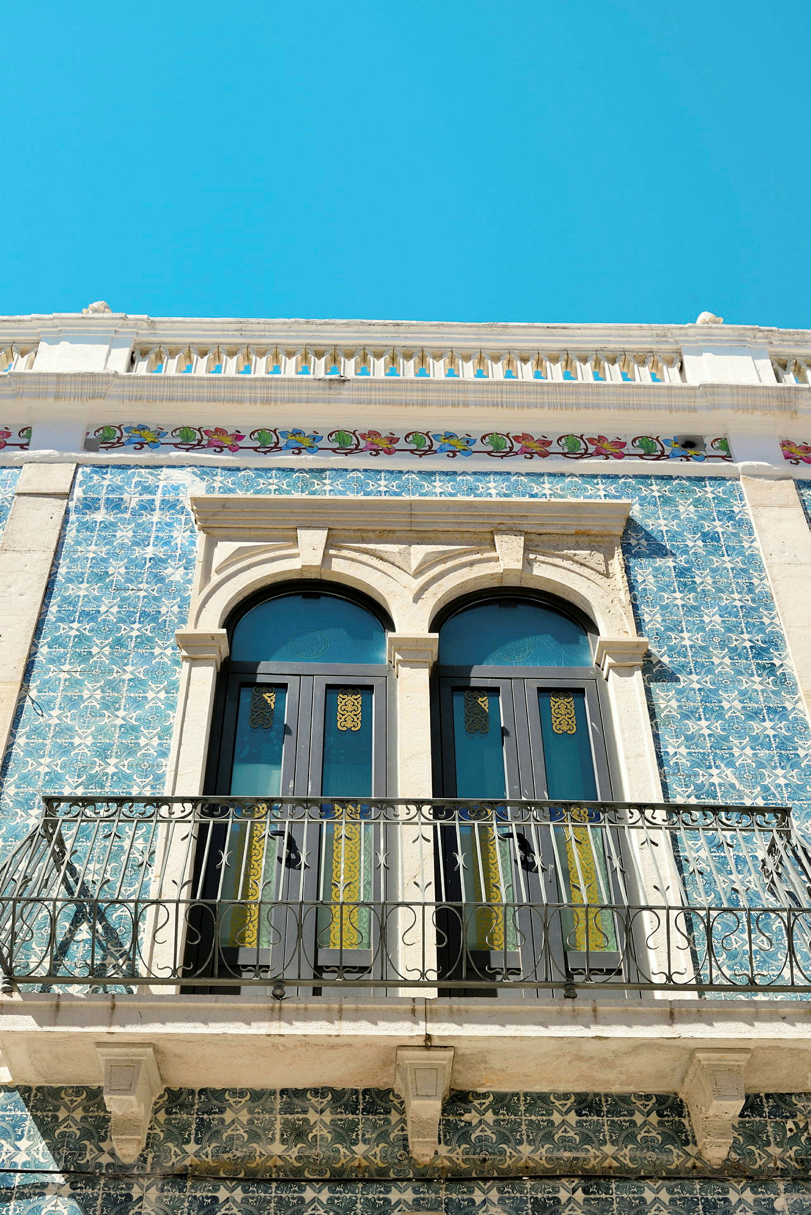 En facade af en bygning med blå mosaikfliser, to buede vinduer og en smedejernsbalkon. (AI-genereret tekst)