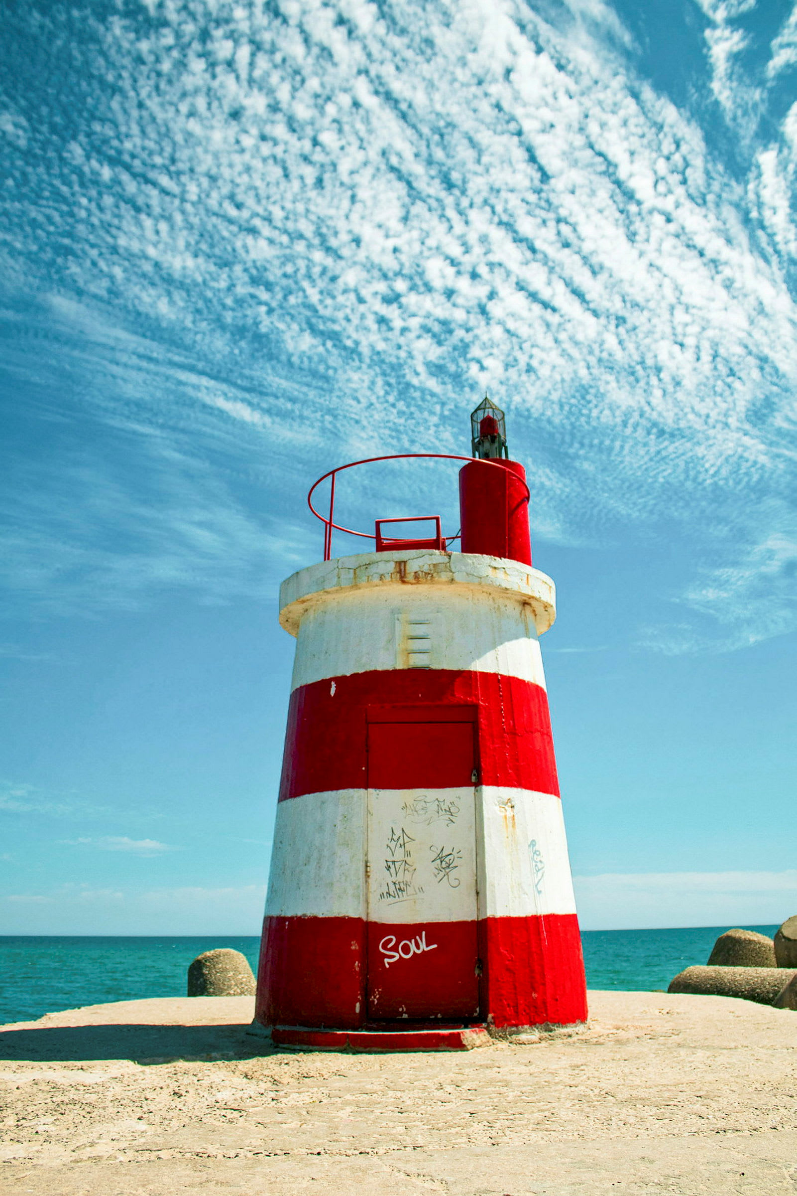 Et fyrtårn med røde og hvide vandrette striber står på en betonbase ved havet med en blå himmel i baggrunden. (AI-genereret tekst)