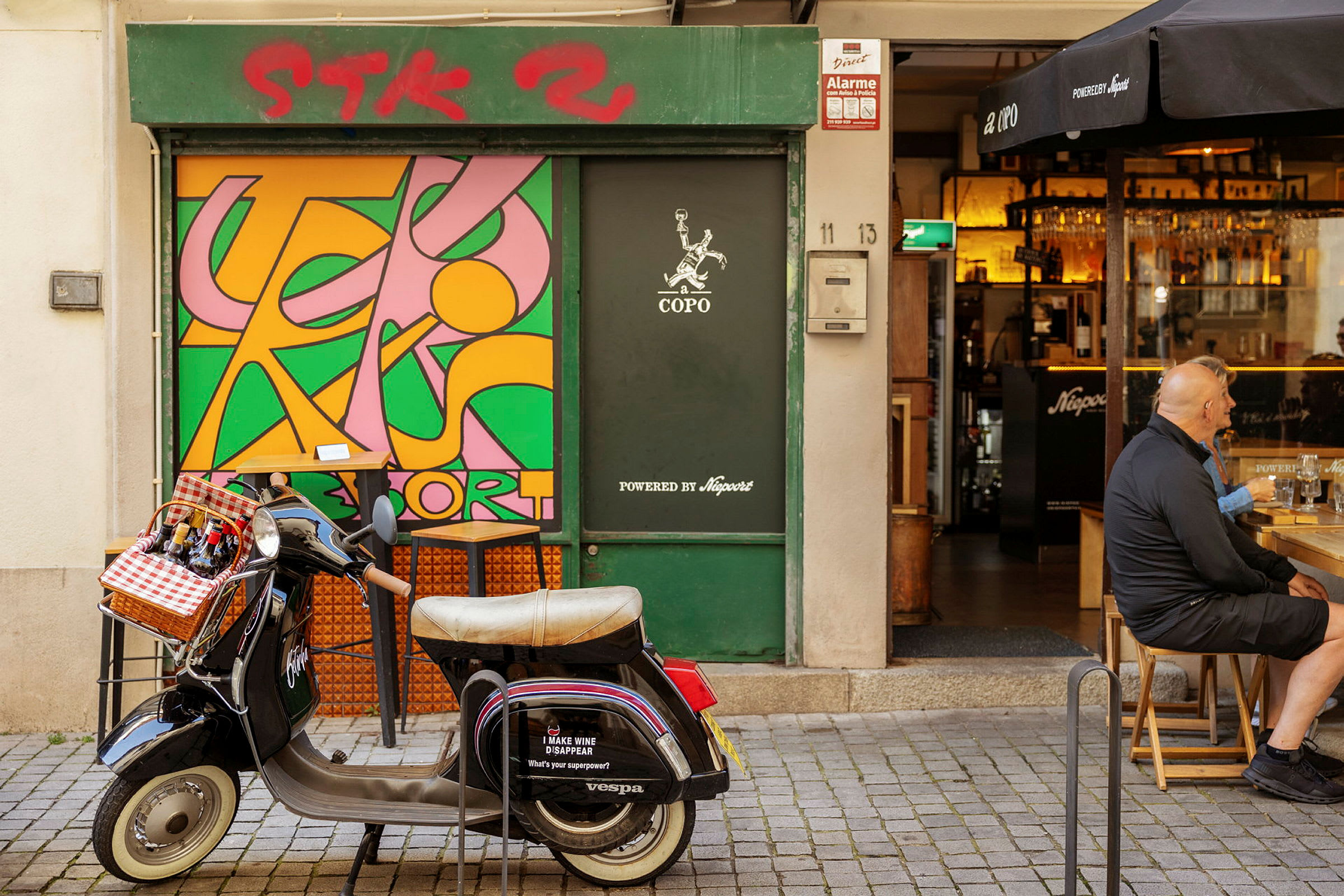 En Vespa med en kurv foran til flasker står parkeret foran en butik med et farverigt vægmaleri. (AI-genereret tekst)