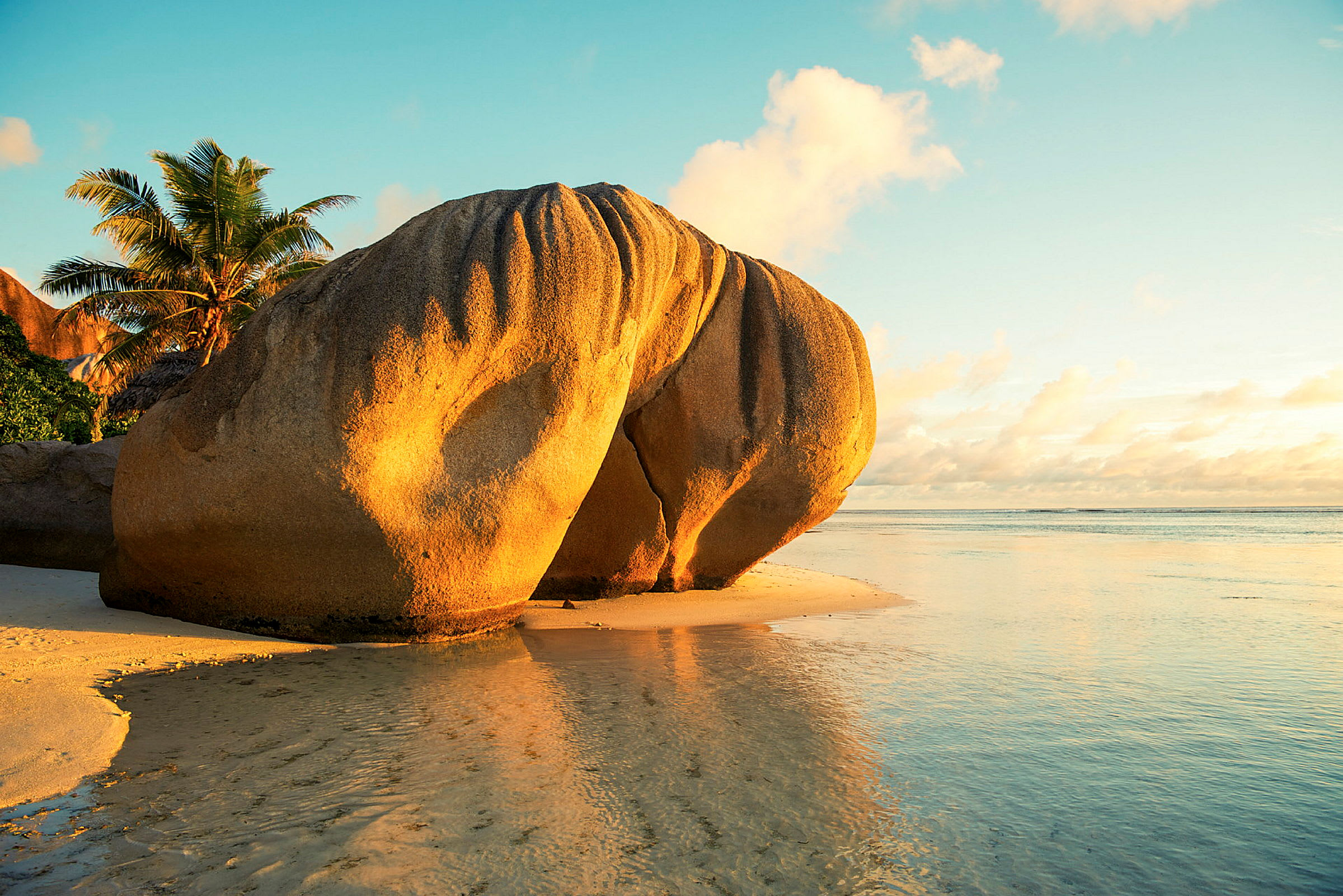 En stor sandstensblok står på stranden med solnedgangen i baggrunden og en palme i baggrunden. (AI-genereret tekst)