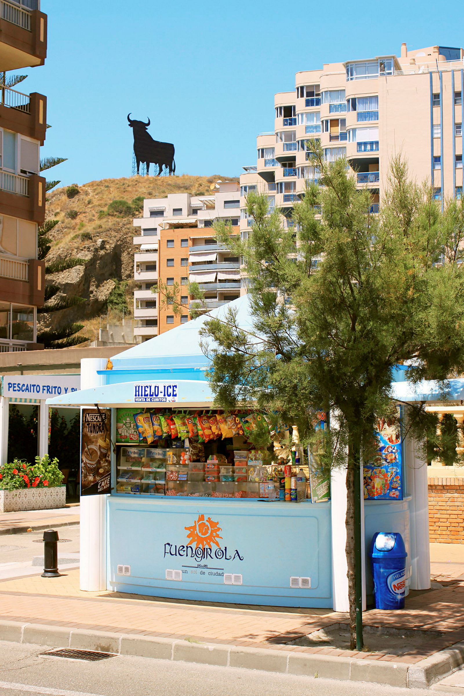 En kiosk omgivet af palmer med bygninger og en tyresilhuet i baggrunden. (AI-genereret tekst)