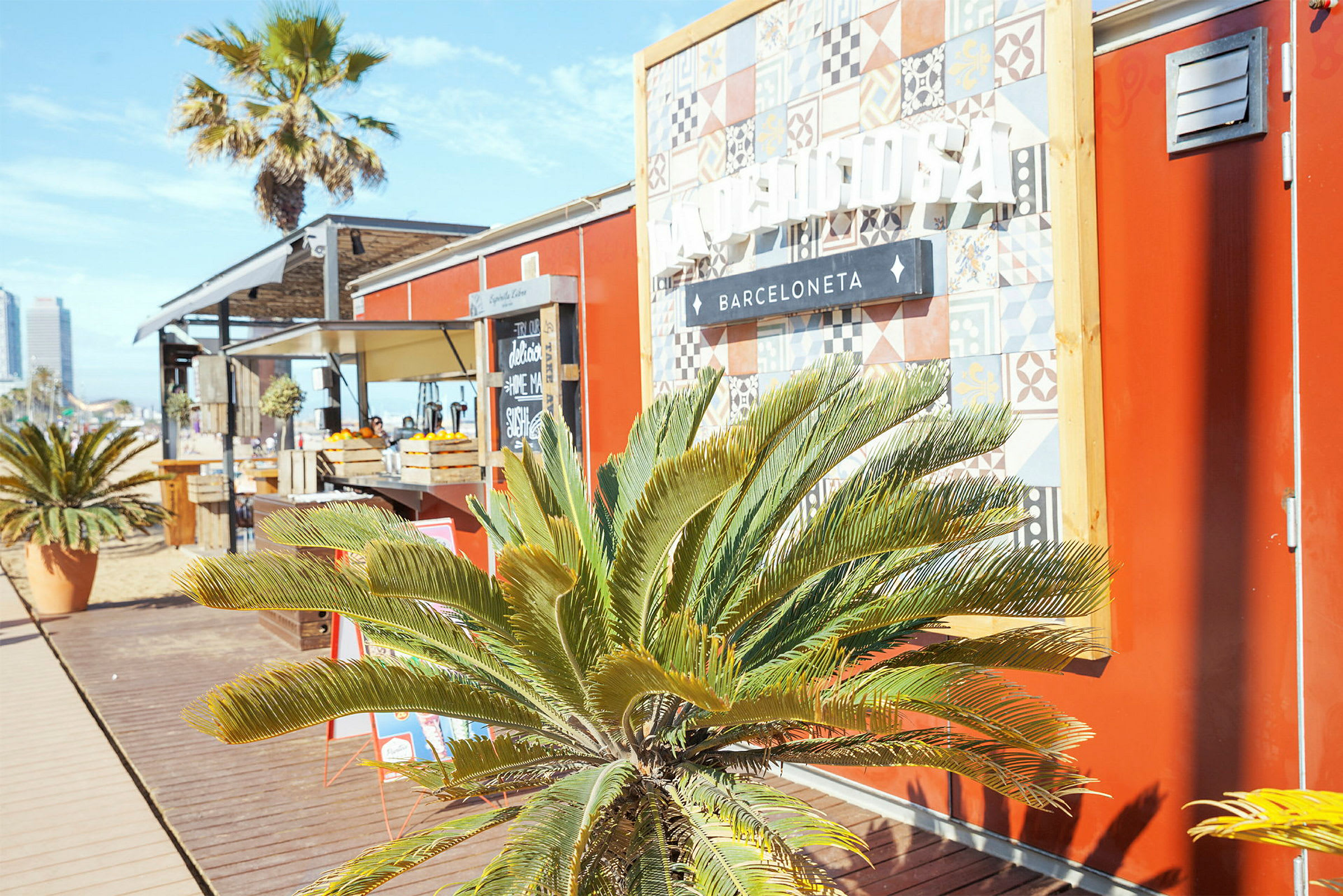 En lille strandbar med røde vægge, et dekorativt skilt og grønne palmeplanter foran. (AI-genereret tekst)