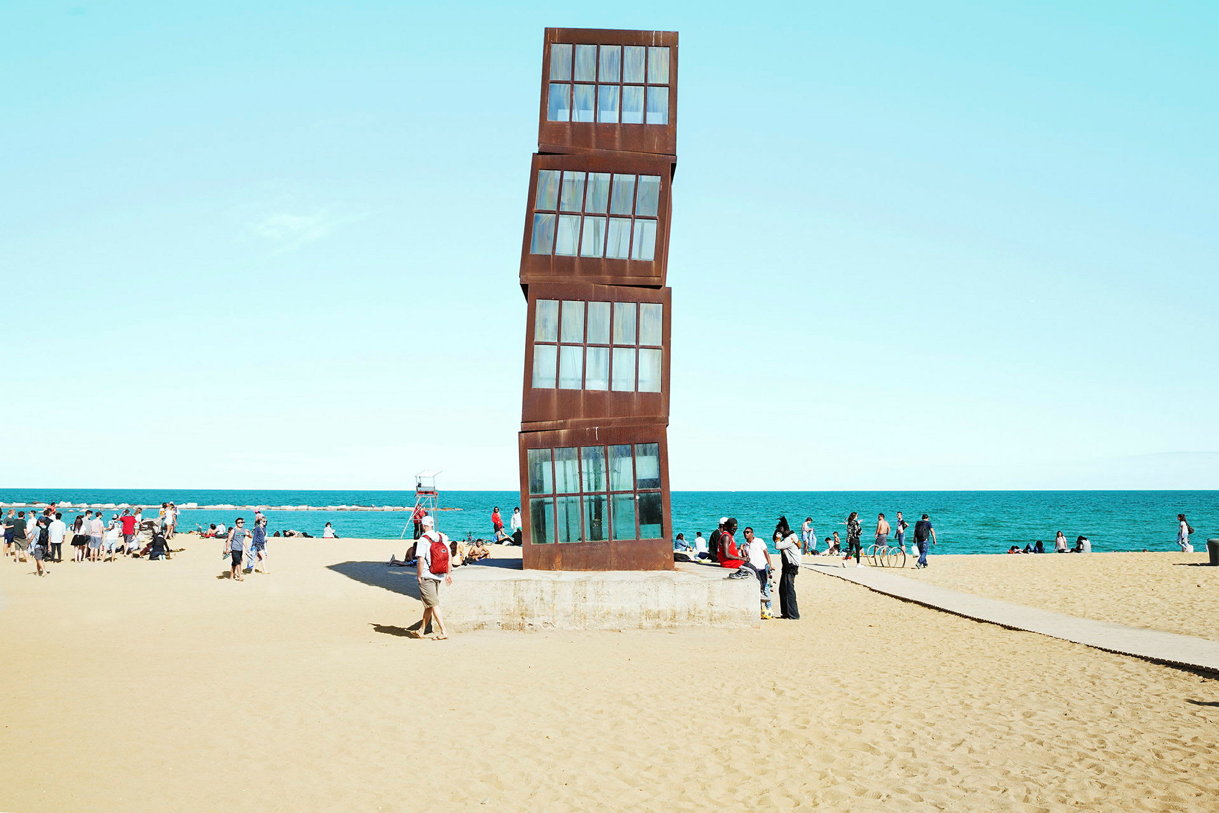 Billede af en strand med en moderne kunstskulptur i fokus samt mennesker i baggrunden. (AI-genereret tekst)