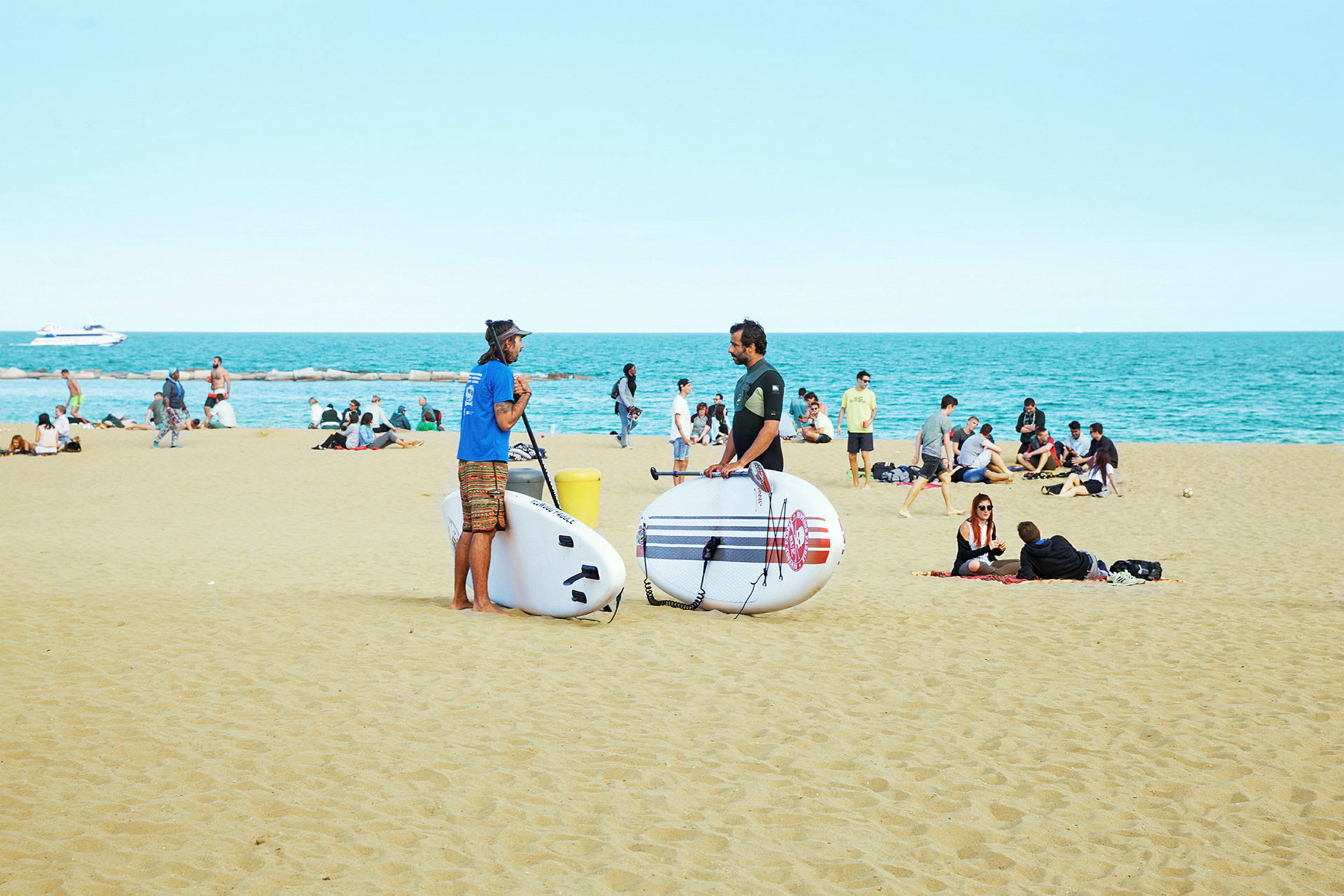 To personer står på en strand med oppustelige paddleboards, mens andre sidder på sanden, og havet ses i baggrunden. (AI-genereret tekst)