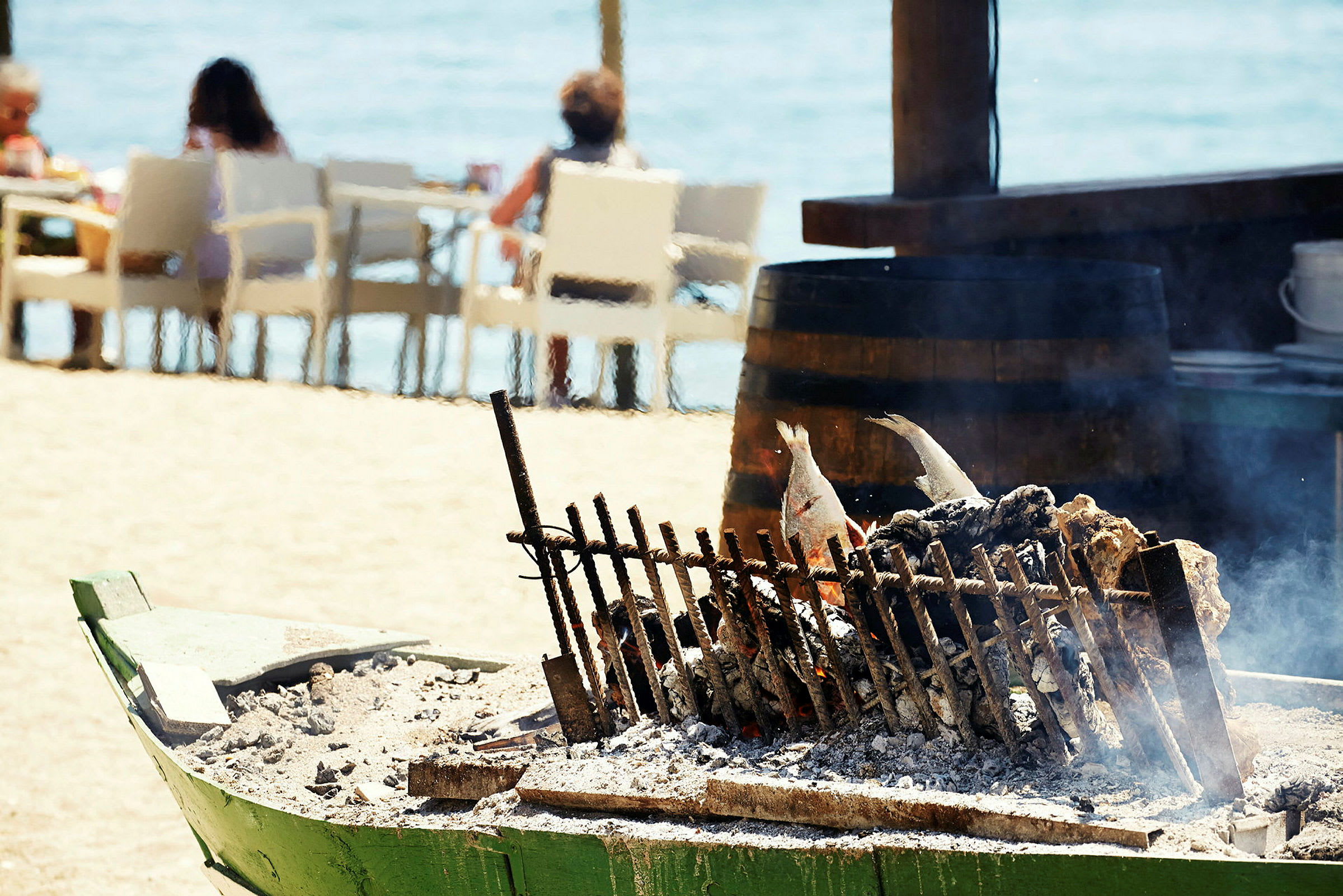 Grillede fisk på et spyd over gløder, med personer, der sidder ved strandborde i baggrunden. (AI-genereret tekst)