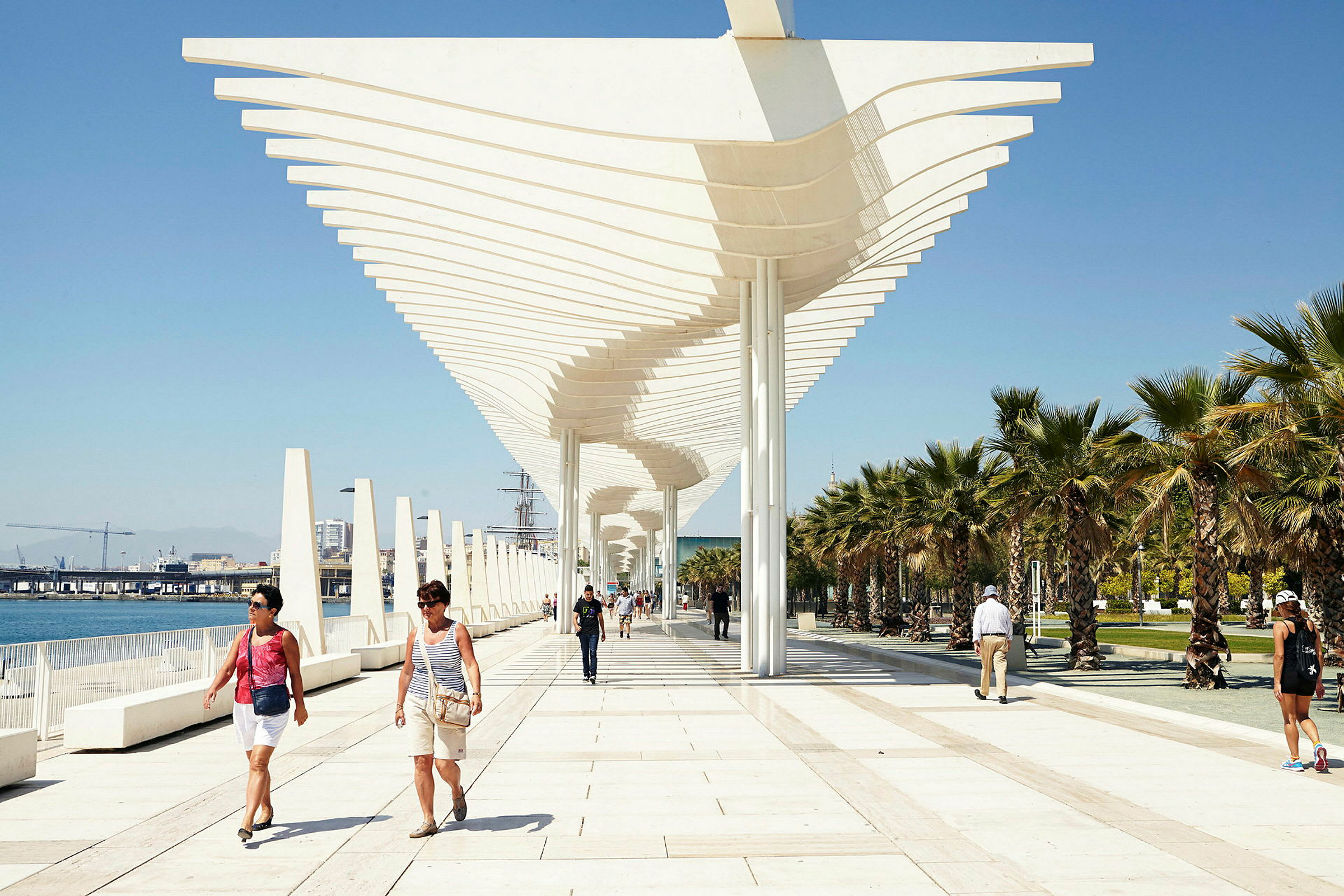 En promenade med moderne arkitektur og palmer på en solrig dag. Folk går langs gangstien. (AI-genereret tekst)