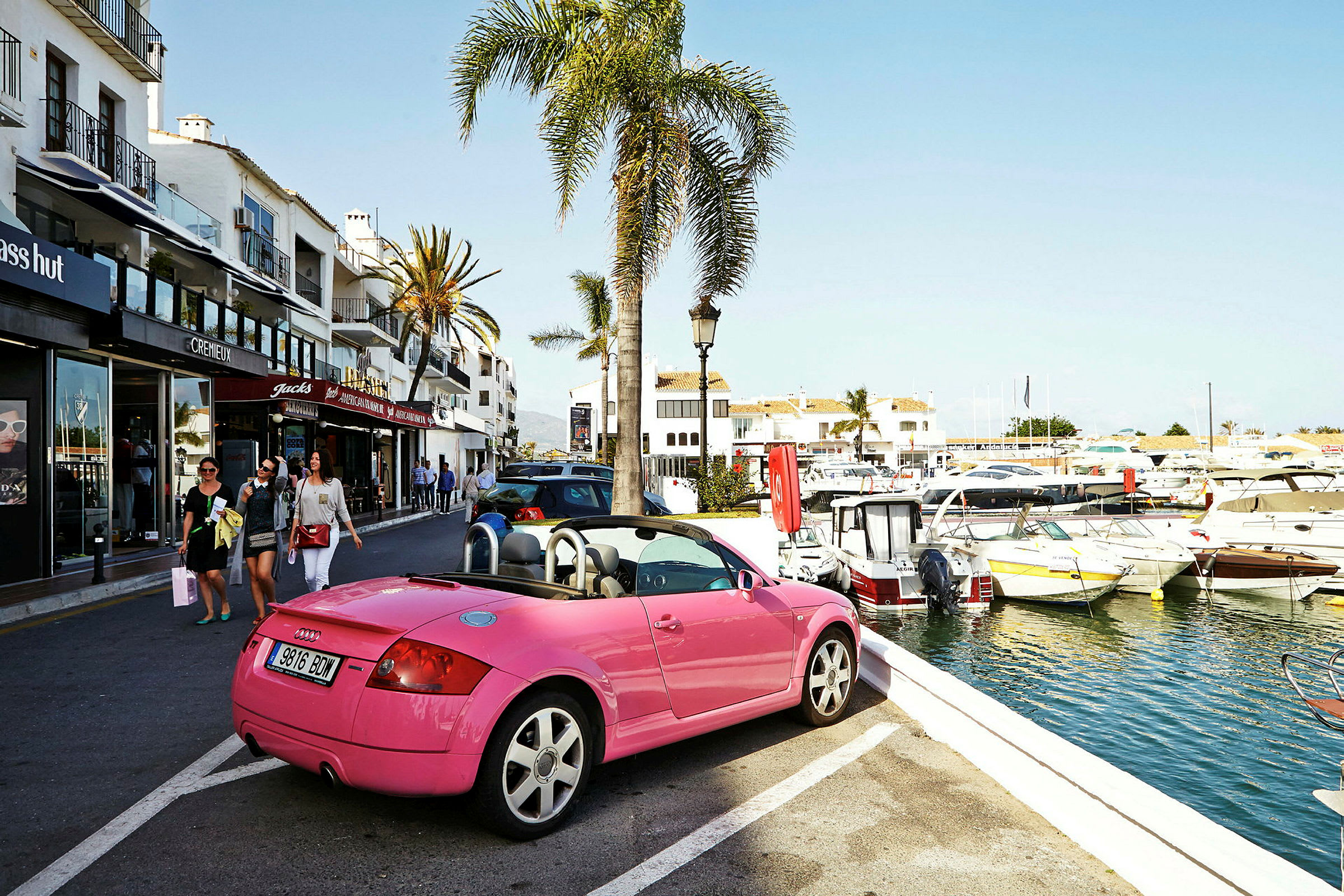 En pink cabriolet parkeret tæt på en havn med både, ved siden af en gade med butikker og palmer. (AI-genereret tekst)
