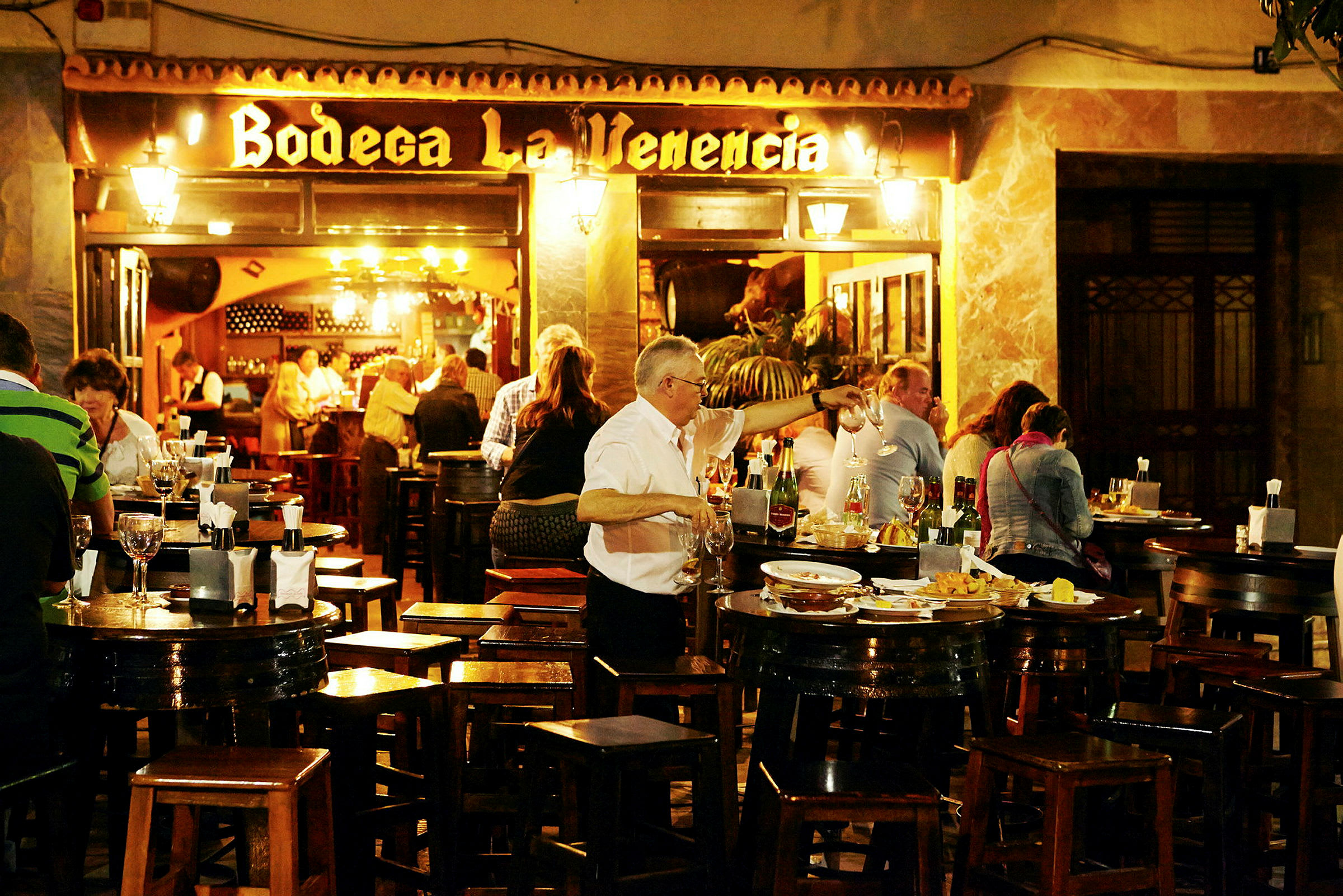En udendørs servering med borde og stole, hvor folk spiser og drikker. Et skilt med teksten 'Bodega La Venencia' ses i baggrunden. (AI-genereret tekst)