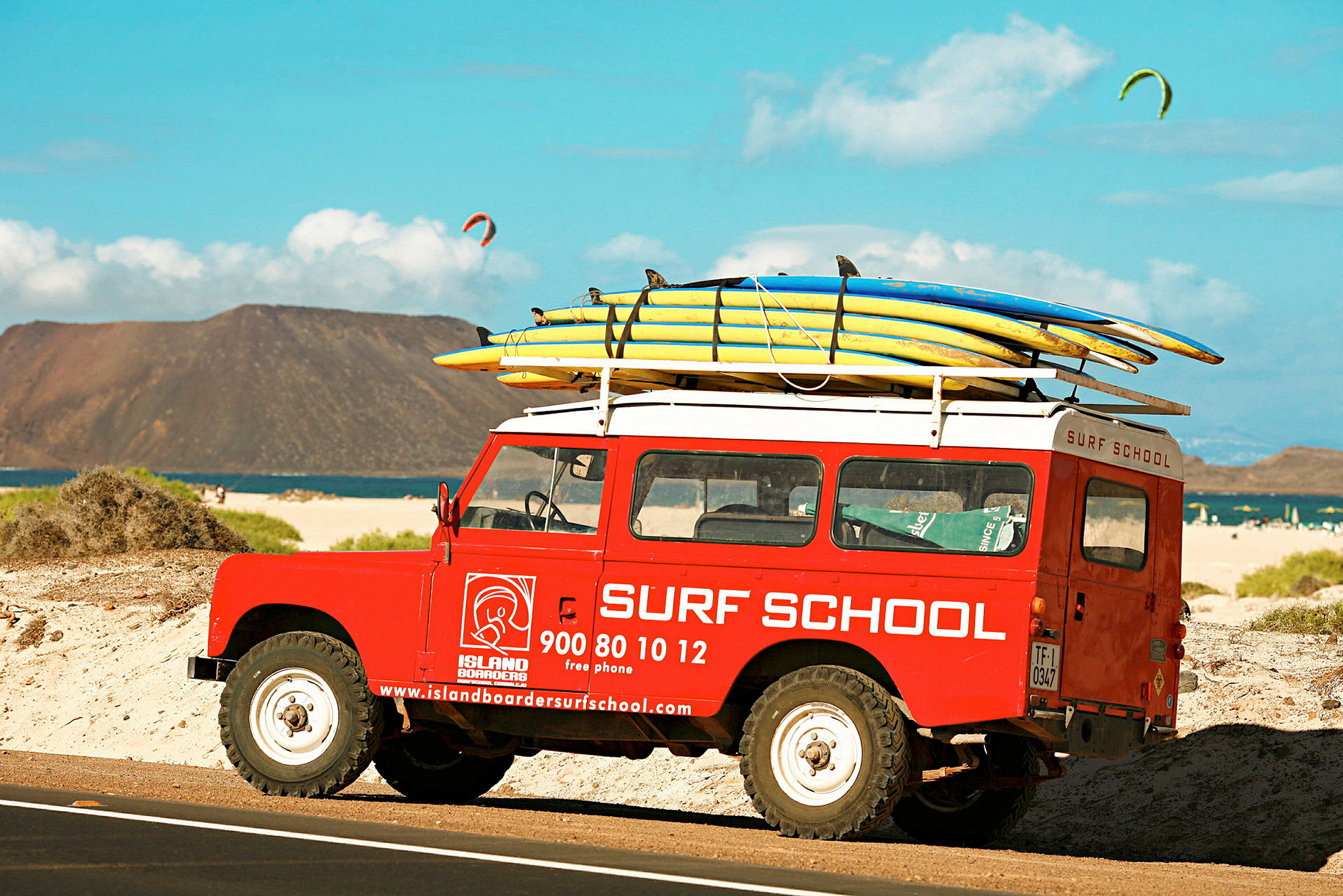 En rød bil med teksten "Surf School" står parkeret på en strand med surfbrætter fastspændt på taget. (AI-genereret tekst)