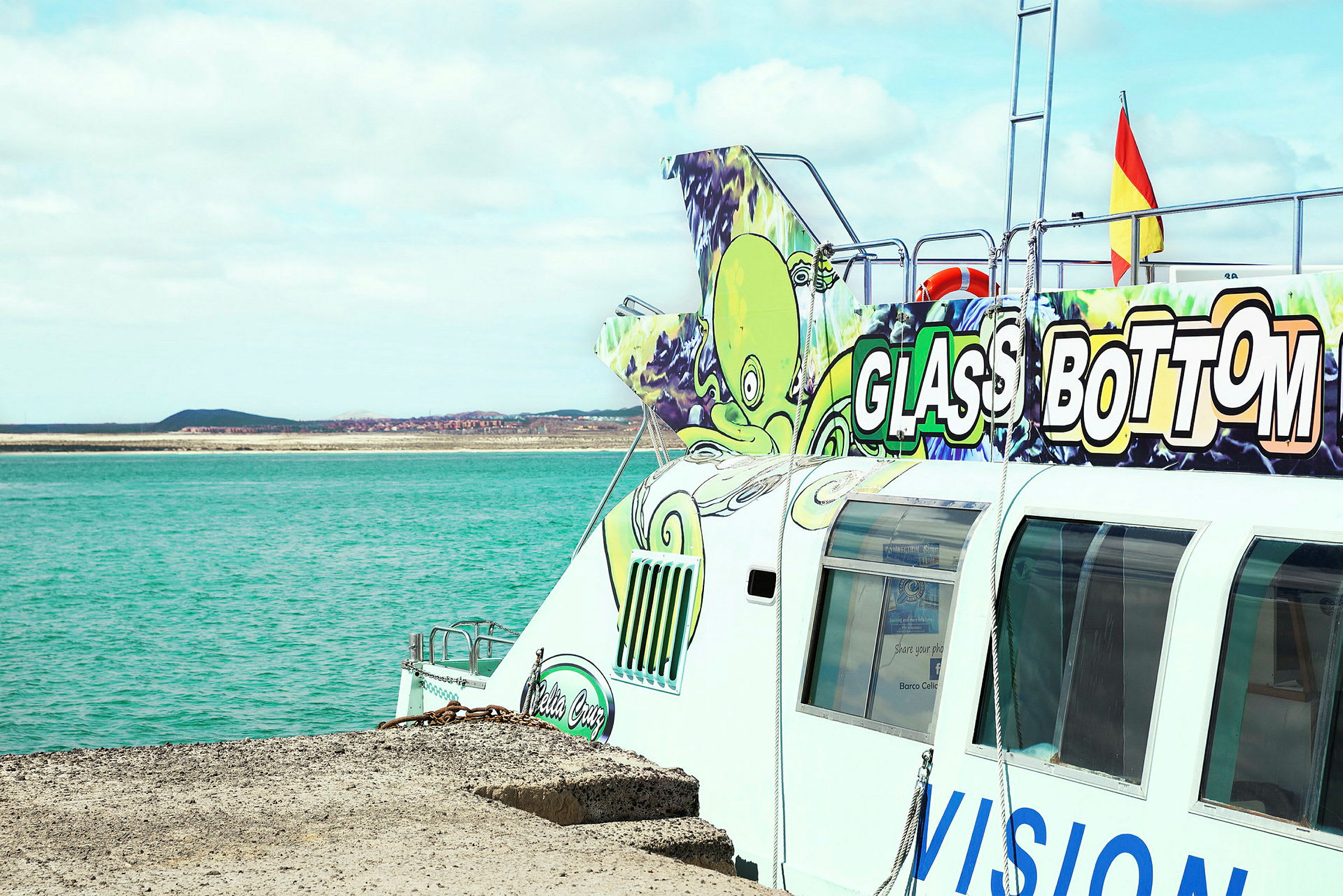 Billede af en glasbundsbåd, som ligger ved en kaj tæt på et turkis hav. Fartøjet har farverige illustrationer, og teksten "Glass Bottom" er klart synlig. (AI-genereret tekst)