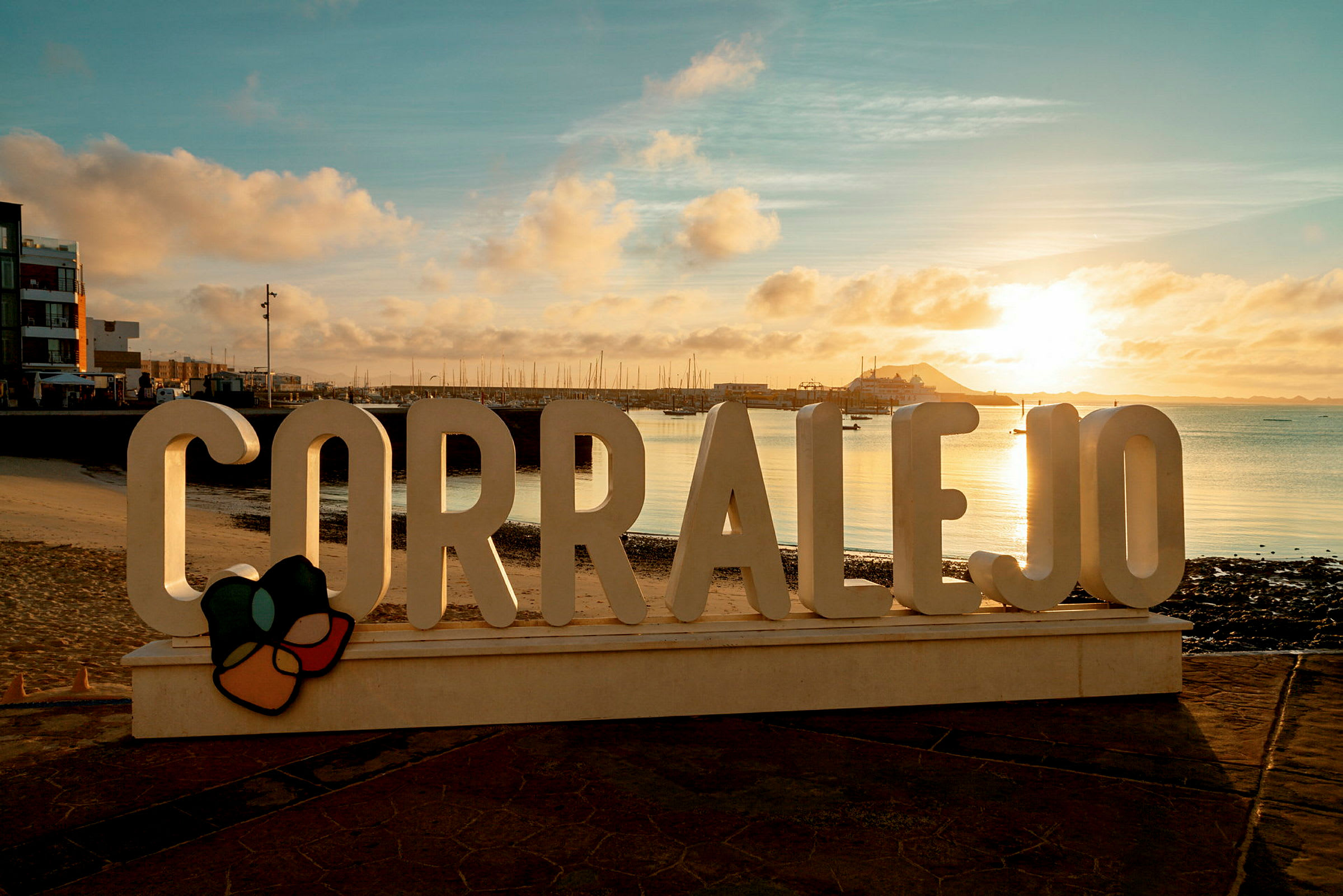 Skiltet 'Corralejo' ved en promenade med havet og en mole i baggrunden under en solnedgang. (AI-genereret tekst)