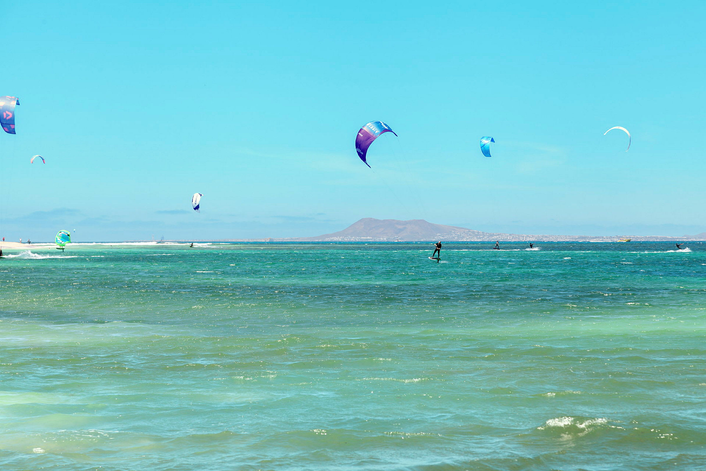 Windsurfere, der sejler på det blå vand med bjerge i baggrunden. (AI-genereret tekst)