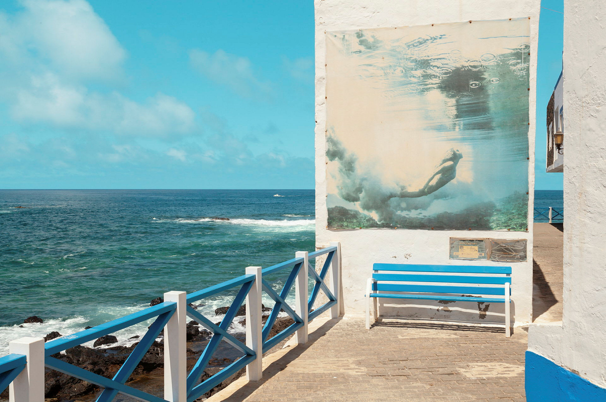 En blå bænk ved siden af et hvidmalet rækværk med udsigt over havet og en væg med en plakat af en dykker. (AI-genereret tekst)
