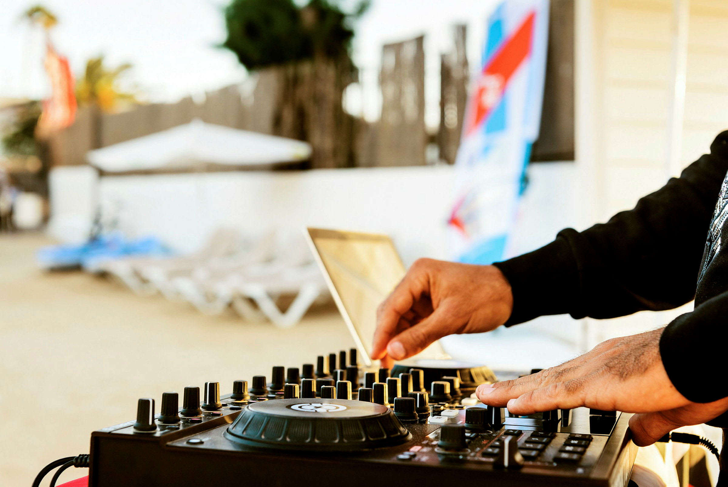 Et nærbillede af en DJ, der justerer knapper på en mixer ved en strand med liggestole i baggrunden. (AI-genereret tekst)