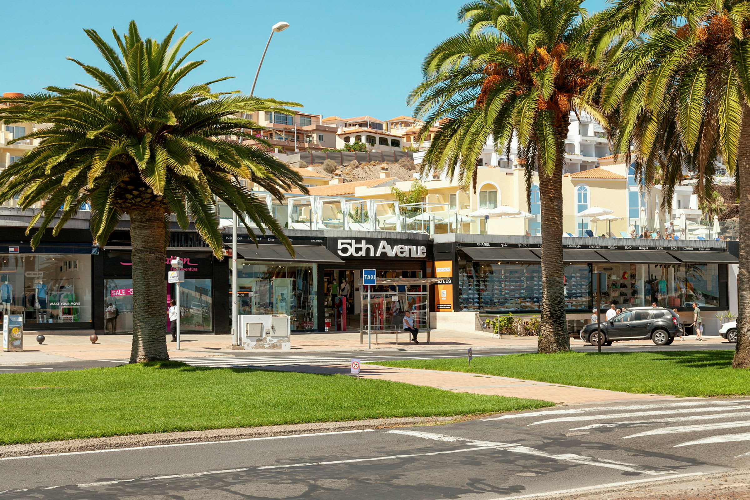 Et shoppingområde med palmer, grønt græs og en butik med skiltet "5th Avenue". (AI-genereret tekst)